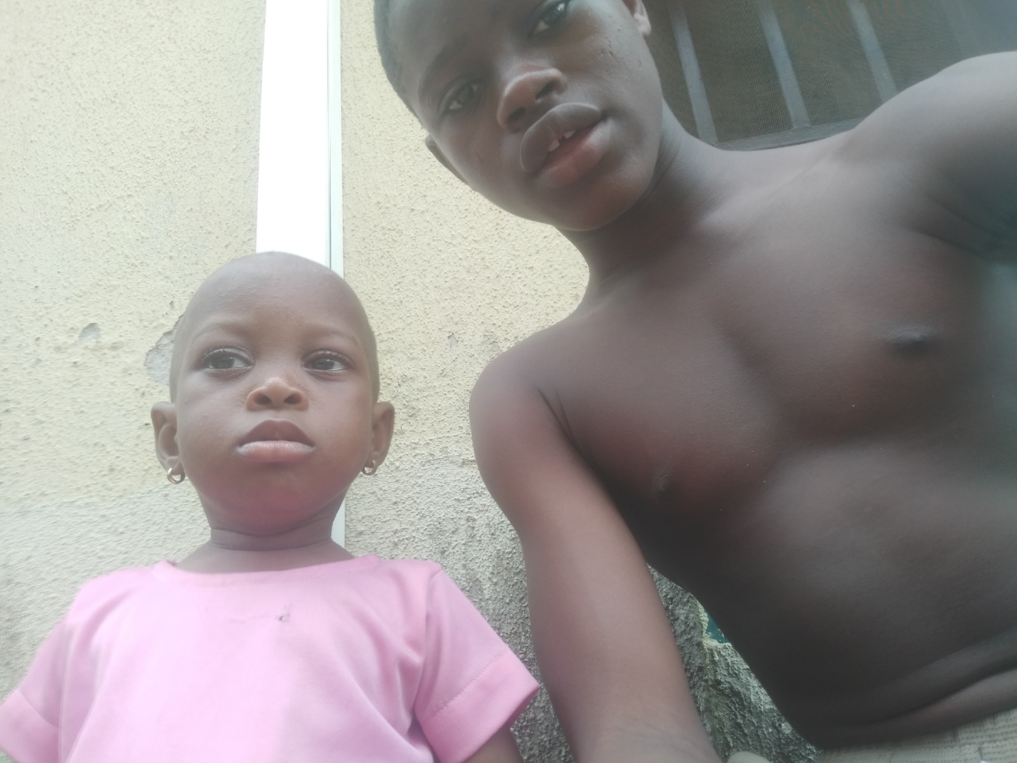 Two young children are in an outdoor setting. One child is wearing a pink shirt and small, round earrings. The other child is shirtless. The background consists of a textured wall and a white vertical structure. The scene appears to be in the city of Shangisha, Nigeria.