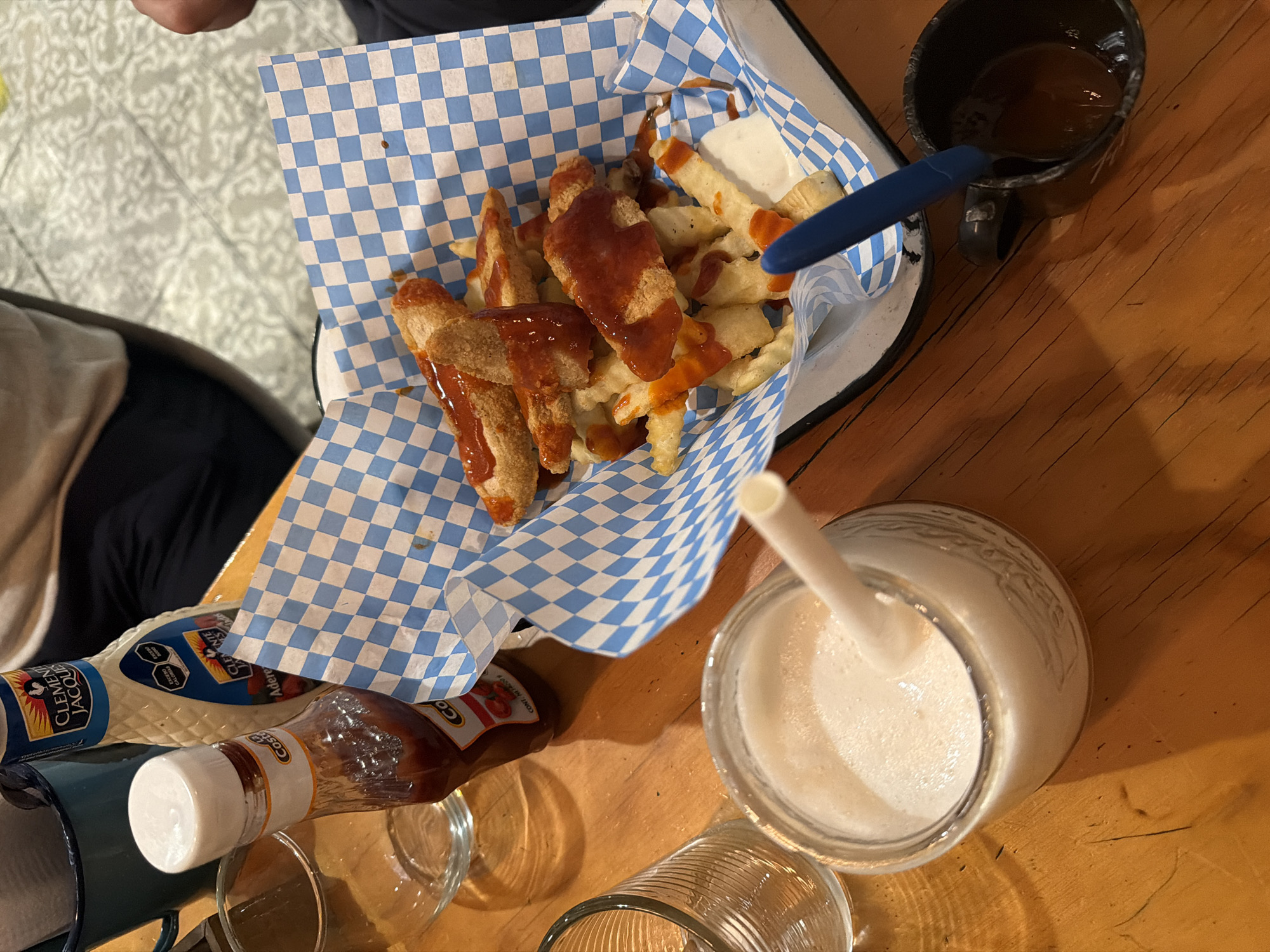 A high-angle shot depicts a wooden table in what appears to be a restaurant setting. The table is populated with a variety of food and drink items. Dominating the scene is a metal basket lined with blue and white checkered paper, containing French fries and what seem to be chicken fingers, both drizzled with a red sauce. Beside the basket, there's a small cup containing a dark liquid, with a blue plastic spoon resting inside. Further down, a milkshake or similar blended drink is visible in a glass jar with a white straw inserted. Condiment bottles, including mayonnaise and ketchup, are present, along with two empty glasses. A person is partially visible in the upper-left corner of the frame. The setting is suggestive of Puebla, Mexico, based on the common use of similar food presentation styles and the types of condiments, although no specific location is identifiable from the image.