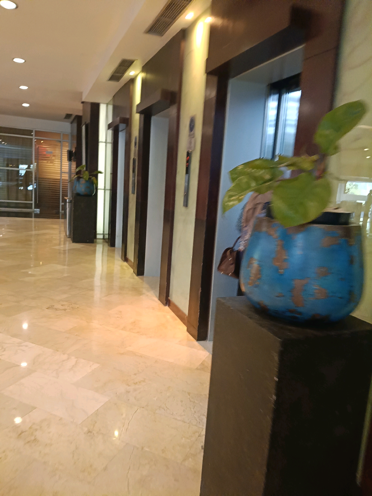 The image depicts an indoor hallway in a building, likely a hotel or office, with a polished marble floor and vertical wooden paneling framing elevator doors. In the foreground, to the right, a large, distressed blue planter holds a green plant. Behind it, a dark pedestal supports the planter. The hallway stretches into the distance, lined with multiple elevator entrances, some of which are illuminated. Through the elevator doors, reflections of the interior and possibly windows can be seen. The lighting appears to be artificial, with recessed ceiling lights contributing to the bright ambiance. The overall impression is one of a clean, modern, and well-maintained public space. Given the context provided, this scene is situated in North Jakarta, Indonesia.