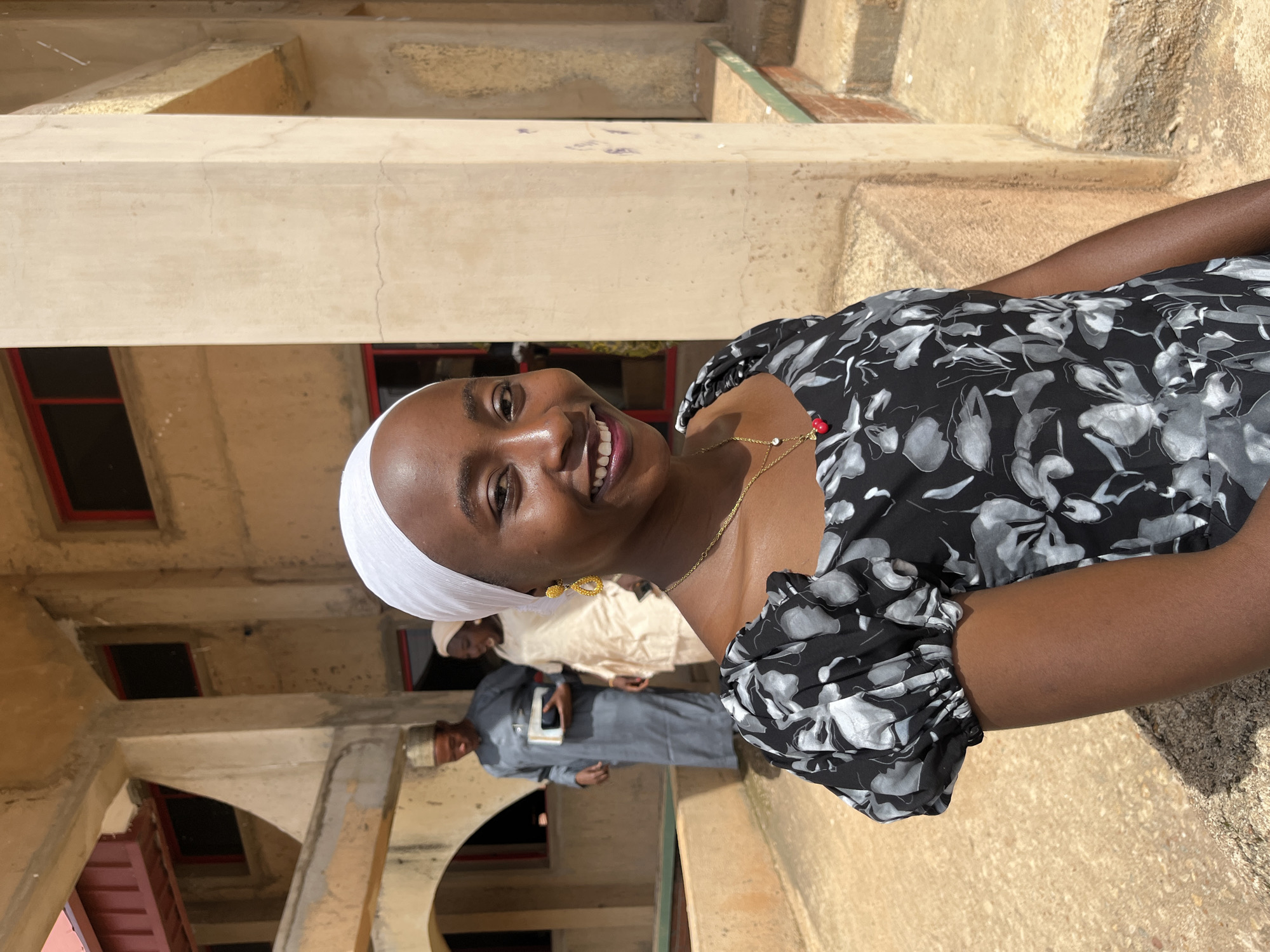 In Bukuru, Nigeria, a young Black woman wearing a white headscarf and a black and white floral dress smiles at the camera. She is adorned with a gold necklace featuring a red pendant and yellow earrings. Behind her, two individuals are visible; a man in a grey traditional outfit holding a book, and a woman in a light-colored outfit. They are standing on a concrete path in front of a light-colored building with red-trimmed windows. The architecture suggests a public or institutional setting.