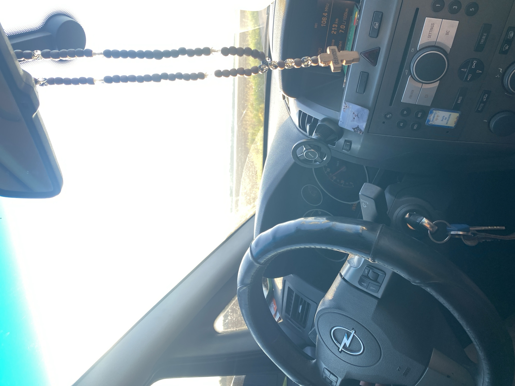 The image shows the interior of a car, looking forward from the driver's perspective. A rosary with a wooden cross hangs from the rearview mirror. The steering wheel, with an Opel logo, is visible on the left. The dashboard includes a radio with a display showing "106.4 MHz" and "213 km." The scene appears to be set during the daytime, with bright sunlight coming through the windshield, suggesting an outdoor environment, possibly a road. The location is specified as the city of Maiske, Ukraine.