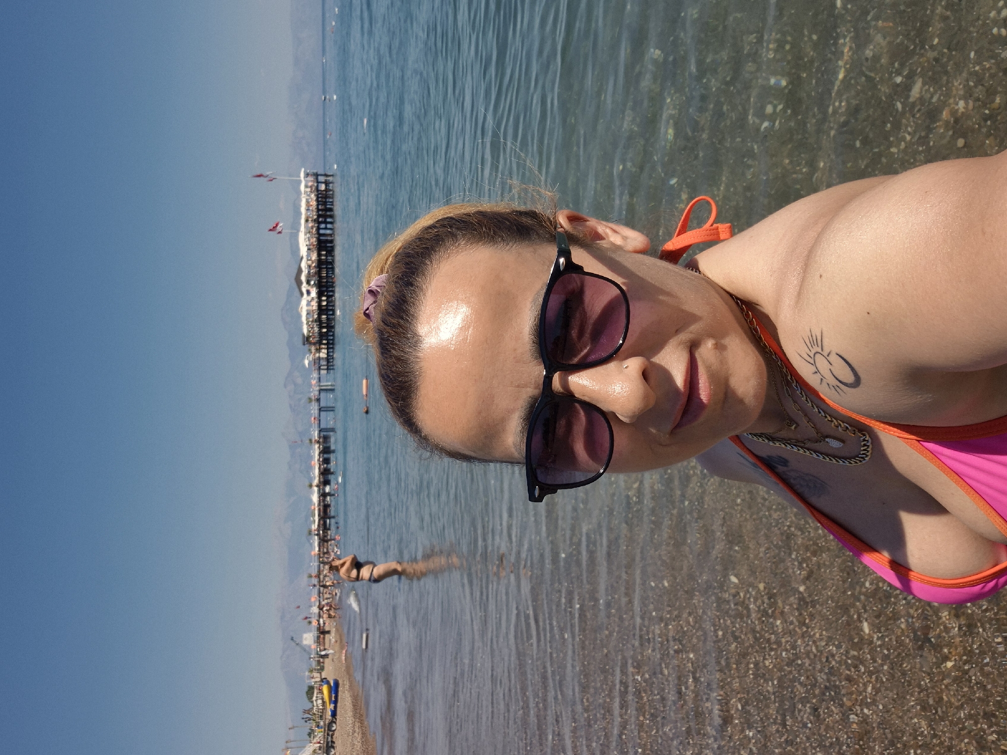 The image is a selfie taken by a woman on a beach. She is wearing a bright pink bikini and sunglasses. The water is clear and shallow, with pebbles visible on the seabed. In the background, there is a pier with people and umbrellas, and a beach with more people and some yellow and blue objects, possibly beach chairs or toys. The sky is clear and blue, suggesting a sunny day. The woman's face is turned towards the camera, and she appears to be smiling or smirking. There is a tattoo of a sun with rays on her upper arm, and she is wearing a gold necklace. The image is oriented vertically, with the horizon line running horizontally across the middle. The overall mood is casual and relaxed, indicative of a vacation or day at the beach.
