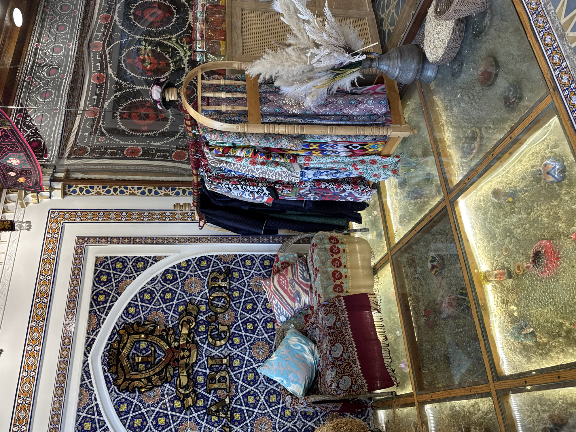 The image shows the interior of a shop in Samarkand City, Uzbekistan. On the wall, there is a decorative blue and white patterned panel with golden Arabic script. In front of it, pillows and textiles rest on a wicker chair. A wooden rack displays colorful textiles under a vase with feathery plants. The shop's floor is glass-paneled and illuminated from below, showing items underneath. The ceiling is covered in ornate fabric with complex designs.