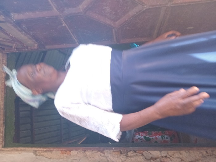 An older individual is captured in what appears to be a vertical, slightly rotated shot, positioned within an opening that resembles a window or doorway. The person is dressed in a white long-sleeved top, possibly with lace detailing on the sleeves, and a dark blue or black long skirt. A light-colored head covering, possibly a scarf or shawl, is visible on their head. Their skin tone is dark. The individual's gaze seems directed slightly upwards and to the left of the frame, with their facial features appearing somewhat blurred.
The setting appears to be a simple, perhaps rustic, structure. The opening they are in has a textured, possibly metallic or wooden frame that looks weathered or aged. Through the opening, behind the person, some patterned fabric (red, white, and green floral/paisley design) can be seen, suggesting an indoor background. Above the person, the ceiling or roof structure is visible, made of corrugated metal or similar material, also showing signs of age.
The lighting is bright, indicating it is daytime. No specific activity beyond standing or looking is clearly discernible. The overall impression is of a casual moment in a simple environment, consistent with locations in rural or less developed areas like Burundu, Kenya. There is no visible text in the image.