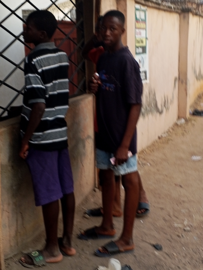 Three young males stand on an unpaved, dusty surface alongside a light beige building facade in Adonkolo, Nigeria. The male on the left, wearing a black and grey horizontally striped short-sleeve shirt, purple shorts, and green-and-black slides, stands in profile, looking left towards a dark metal security grill covering a window or doorway. A dark red door or frame is visible behind the grill. The male in the center, wearing a dark blue short-sleeve t-shirt with a light-colored logo, light blue denim shorts, and dark slides, faces forward, holding a white rectangular object in his right hand and folded pink currency notes in his left hand. A third male, partially visible behind the central figure and to his right, wears a red short-sleeve shirt and dark slides, leaning against the light beige wall. This wall features several affixed vertical posters with text and imagery. Scattered debris is present on the ground.