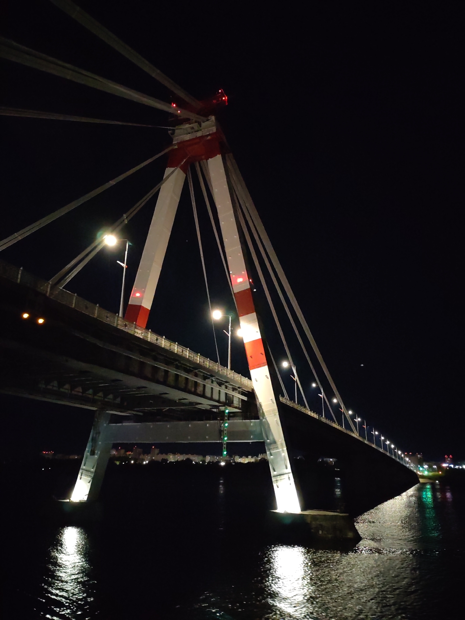 The image depicts a bridge at night, likely the Oktyabrsky Bridge in Cherepovets, Russia. The bridge has a distinctive pylon structure with red and white markings. Cables extend from the top of the pylon to support the bridge deck. Streetlights line the bridge. Water reflects the bridge lights and the cityscape in the background. The scene is dark with visible light sources defining the structure and reflections on the water.