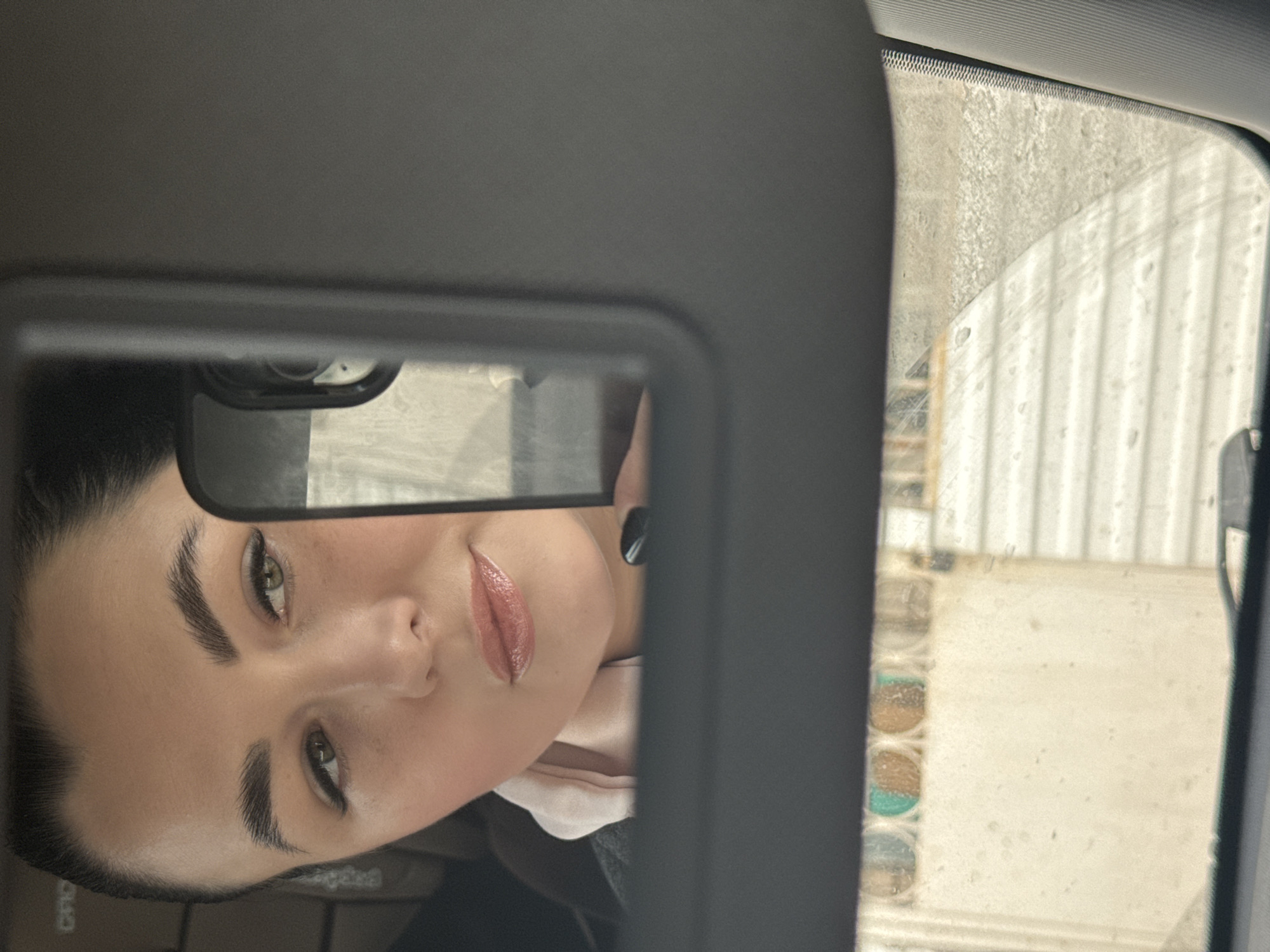 A woman's face is visible in the reflection of a vanity mirror inside a car. She has dark hair, light skin, and is wearing lip gloss. The car's sun visor frames the mirror. Through the car window, a building with corrugated metal siding is visible. The environment outside is cloudy or snowy. It is not possible to determine the location in the city of Chaikovsky, Russia.