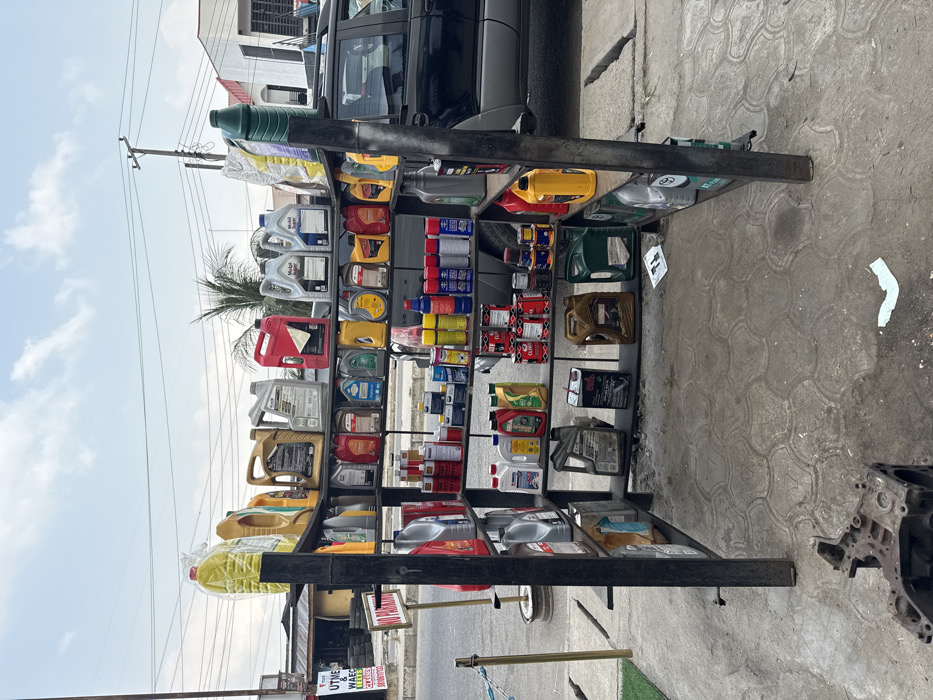 The image depicts an outdoor retail display of automotive products situated on a concrete surface in Badore, Nigeria. A prominent four-tiered, black metal shelving unit, positioned centrally, holds numerous plastic containers of engine oil, transmission fluid, and other automotive lubricants in various colors (yellow, red, silver, black, blue, green). Brands such as Mobil Super are visible on some containers. Cans of spray products and smaller metal cans are also arranged on the lower shelves.
In the immediate foreground to the lower left, a discarded engine block or a similar heavy metallic automotive component rests on the textured concrete ground, which shows minor debris including small pieces of white paper.
To the left of the display, a paved street is visible, bordered by a white line. A faded "NO PARKING" sign is attached to a pole, and another sign, partially obscured, indicates "UTME & WAEC". A stack of black tires is visible behind these signs, adjacent to a low-rise building with a brown roof. A metal stanchion with a blue rope barrier is present near the street.
In the background to the right, a dark-colored SUV, likely a Subaru Forester model, is parked behind the shelving unit, with its left side and roof rack visible. Beyond the vehicle, a light-colored building with a green-trimmed roof is present. Overhead utility lines and poles crisscross against a partly cloudy, light blue sky. A palm tree is visible behind the shelving unit in the mid-distance. The scene is devoid of any visible people or ongoing activity.