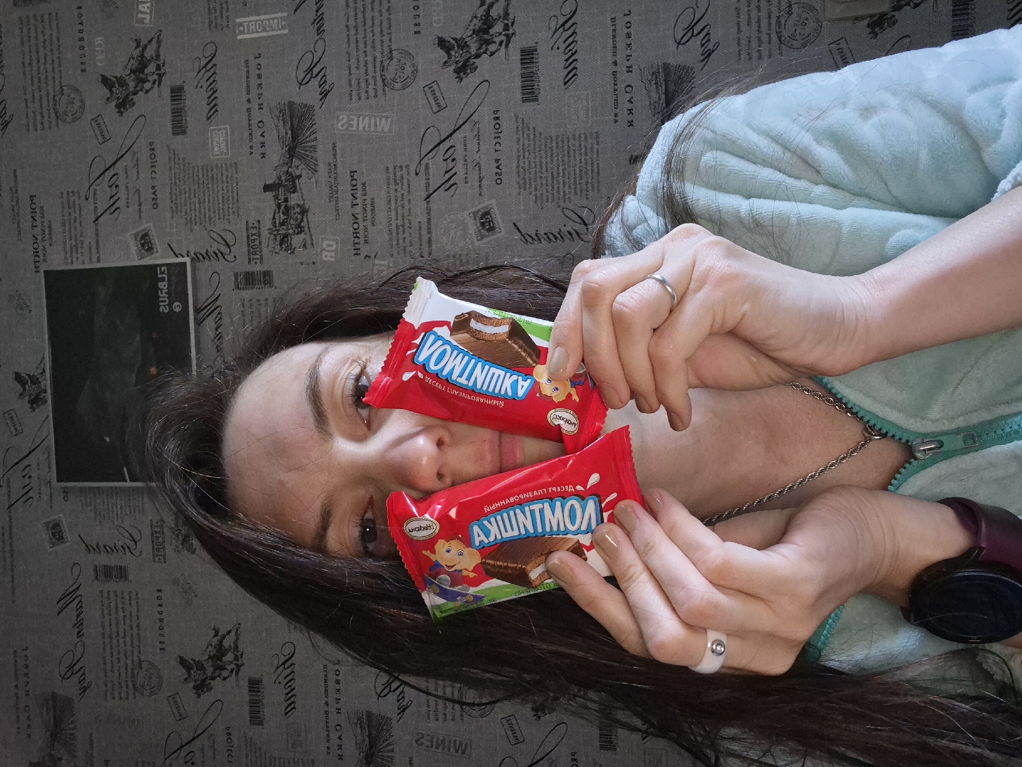 A woman is positioned in front of a patterned wallpaper that simulates a newspaper print. She is lying down, partially covered by a light blue fuzzy garment. Two chocolate bars, branded "Vowintmky", are held in her hands close to her face. She wears two rings, one silver on her right hand, and a white ring on her left. A silver chain necklace adorns her neck. Black headphones are partially visible on her right wrist.