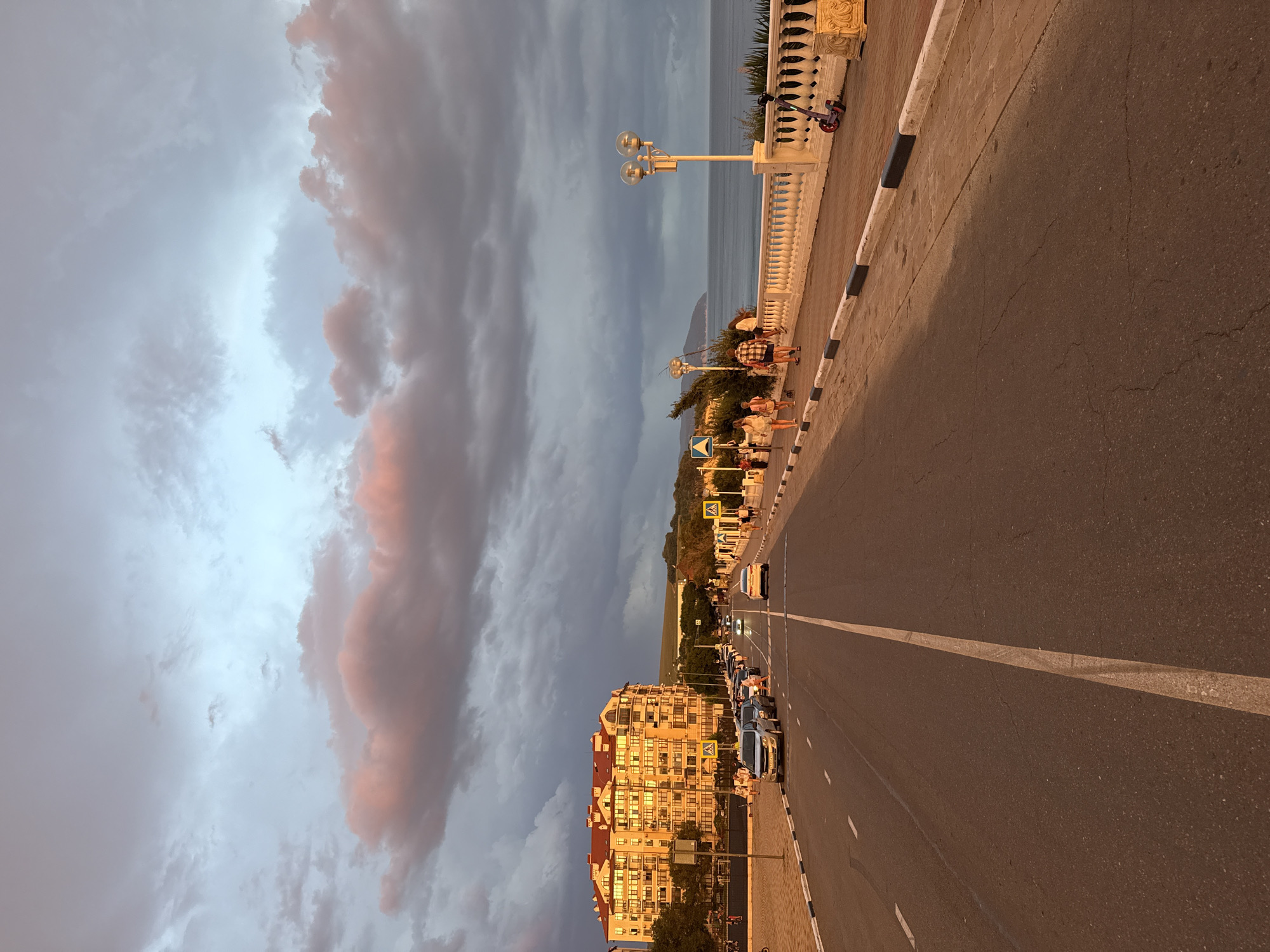 The image captures a street scene in Gelendzhik, Russia, during what appears to be either sunrise or sunset, given the warm, golden light illuminating the scene and the dramatic clouds overhead. The viewpoint is from the middle of a paved road, looking forward.
On the left side of the road, a multi-story building with a red-tiled roof and numerous balconies is prominently featured, catching the strong sunlight. The building is set back slightly from the road, with a paved area and some trees in front of it. Cars are parked along the side of the road.
The road itself is a two-lane asphalt street with a white dividing line. Several cars are driving or stopped on the road, creating a moderate traffic flow. Pedestrians are visible on the right side of the road, walking along a wide sidewalk. This sidewalk is separated from the road by a decorative white balustrade. Streetlights with globe-shaped lamps are spaced along the sidewalk. Further down the sidewalk, a child's scooter is parked.
The environment suggests a coastal city, with the sea visible in the background beyond the sidewalk. The sea is calm, reflecting the sky. The sky is filled with large, dramatic clouds, some tinged with pink and orange from the low-angle sunlight, contrasting with darker, grayer cloud formations. The overall atmosphere is serene, with a sense of urban life by the sea during a picturesque time of day.