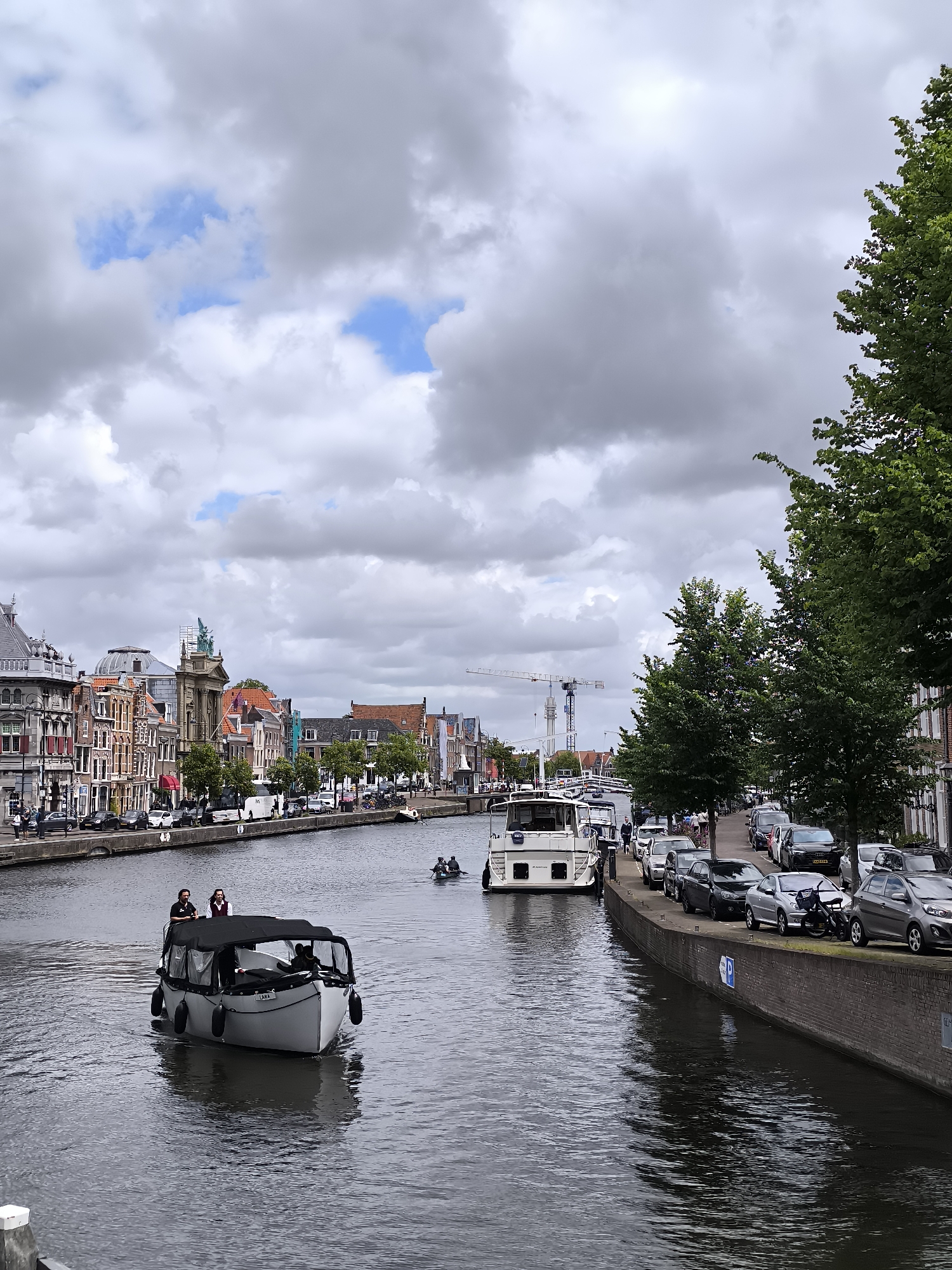 This is a medium shot, taken from a slightly elevated perspective, of a canal in Haarlem, Netherlands. On the left bank of the canal, a row of old, multi-story buildings with ornate facades line the water. Several people are walking on the sidewalk in front of these buildings. A small tour boat with a black canopy is moving down the canal, carrying at least three people. On the right bank, a tree-lined promenade runs parallel to the canal, with a street behind it. Cars are parked along this street. In the background, several cranes are visible, suggesting ongoing construction or development. The sky is overcast with dramatic clouds.