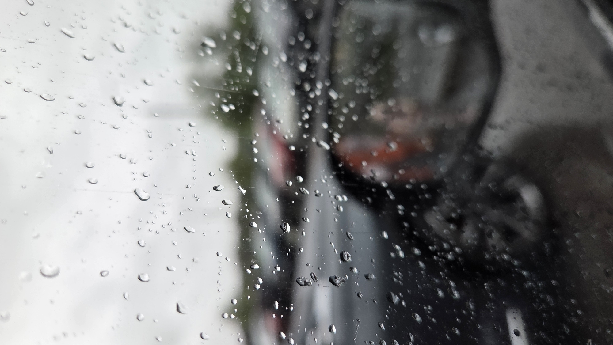 The image is a close-up shot of a car window covered in raindrops. Through the wet glass, a blurred outdoor scene is visible, suggesting a parking lot or street. Several vehicles, primarily in dark colors, are parked in the background, with some hints of red and lighter colored cars. The sky appears overcast and grey. The image captures a typical rainy day scene, possibly in a suburban or urban environment, though specific Saint Petersburg landmarks are not discernible. The focus is on the texture and pattern of the raindrops on the glass, creating a sense of depth and obscuring the details of the outside world. No people are directly visible, and the focus is on the atmospheric effect of the rain.