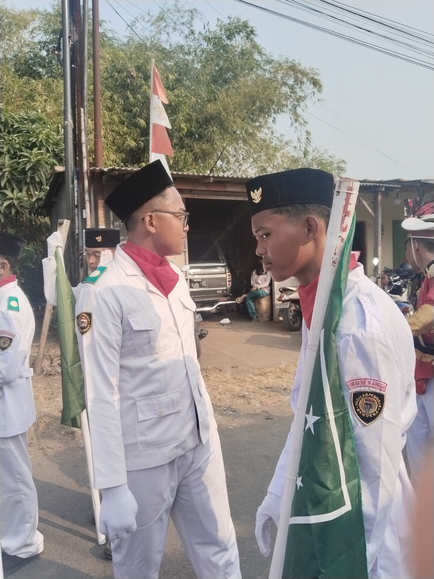 Two young males in white uniforms and black hats stand facing each other outdoors. The male on the left wears glasses and a red scarf around his neck. He holds a green flag with white stars on a pole. The male on the right also wears a red scarf and is holding a flag pole. Both are wearing white jackets with emblems on the left breast. In the background, there is a building with a corrugated metal roof, some trees, and a parked car. The scene appears to be in Margasari, Indonesia, as indicated by the emblem on the uniform.