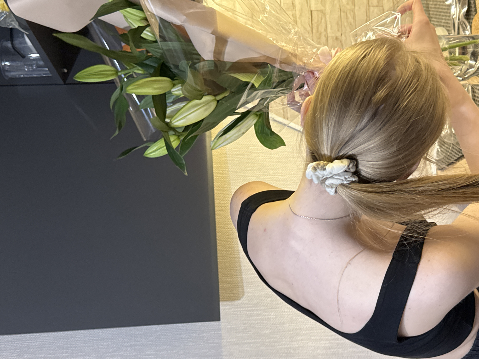 A woman with blonde hair pulled back in a ponytail with a white scrunchie is holding a bouquet of lilies wrapped in clear plastic. She is wearing a black sports bra and a thin silver necklace. The bouquet is partially over a dark gray object, possibly a table or cabinet. The background shows a neutral-colored wall with a textured surface. The location is possibly Perm, Russia.
