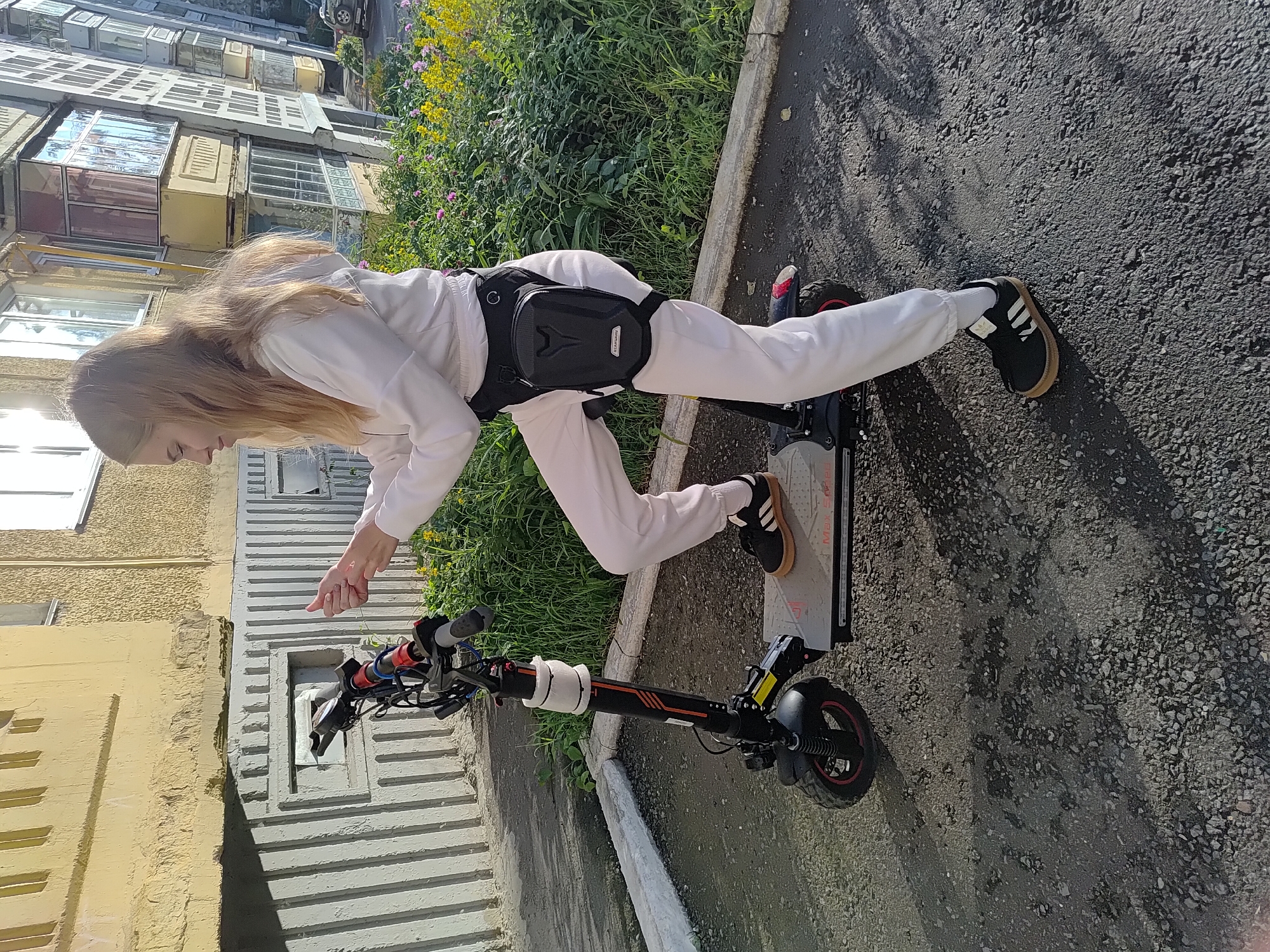 A young woman with long blonde hair is on an electric scooter outdoors. She is wearing a white hoodie and white pants, with a black fanny pack around her waist. Her left leg is extended, touching the ground, while her right foot is on the scooter's deck. The scooter is black with red accents and has a digital display on the handlebars. The woman is looking down and appears focused.
The scene is a paved area next to a building with a textured yellow facade and multiple windows. A patch of green grass and wildflowers is visible to the left of the scooter. The ground is covered in small gravel. Sunlight casts shadows on the ground, suggesting it is daytime. The building's architecture and the presence of flowering plants hint at a residential area in Saransk, Russia. The woman's posture and the scooter suggest she might be practicing or preparing to ride.