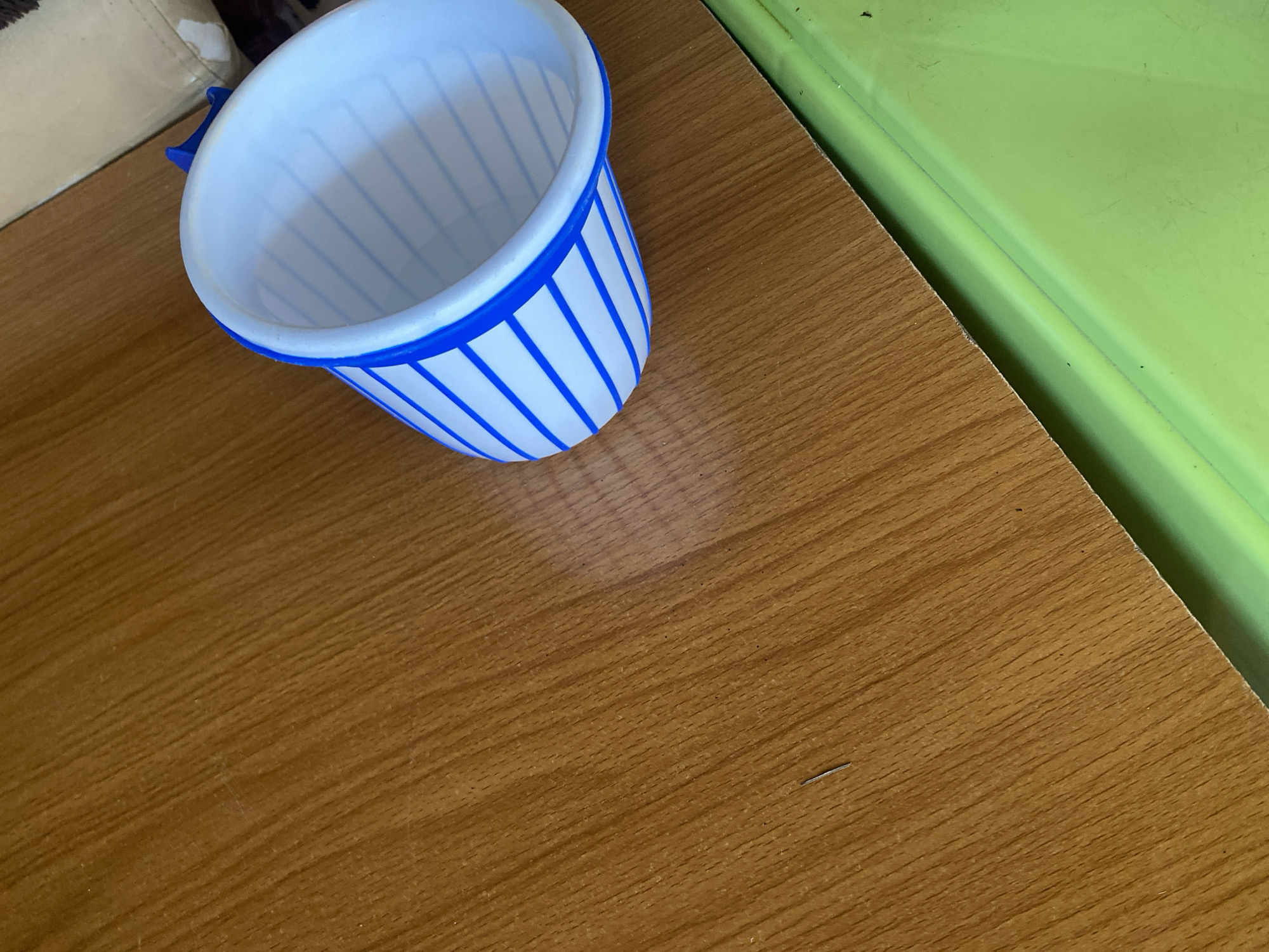 A white plastic cup with blue stripes is placed on a wooden surface. The cup is oriented towards the right side of the frame and appears to be empty. The wooden surface has a visible wood grain pattern. In the background, to the right of the cup, a portion of a light-colored fabric object is visible, possibly furniture. Below the wooden surface, a bright green surface is partially visible. There are no people or discernible activities taking place. The scene depicts an indoor setting with everyday objects. The image location is Jauro Yinu, Nigeria.
