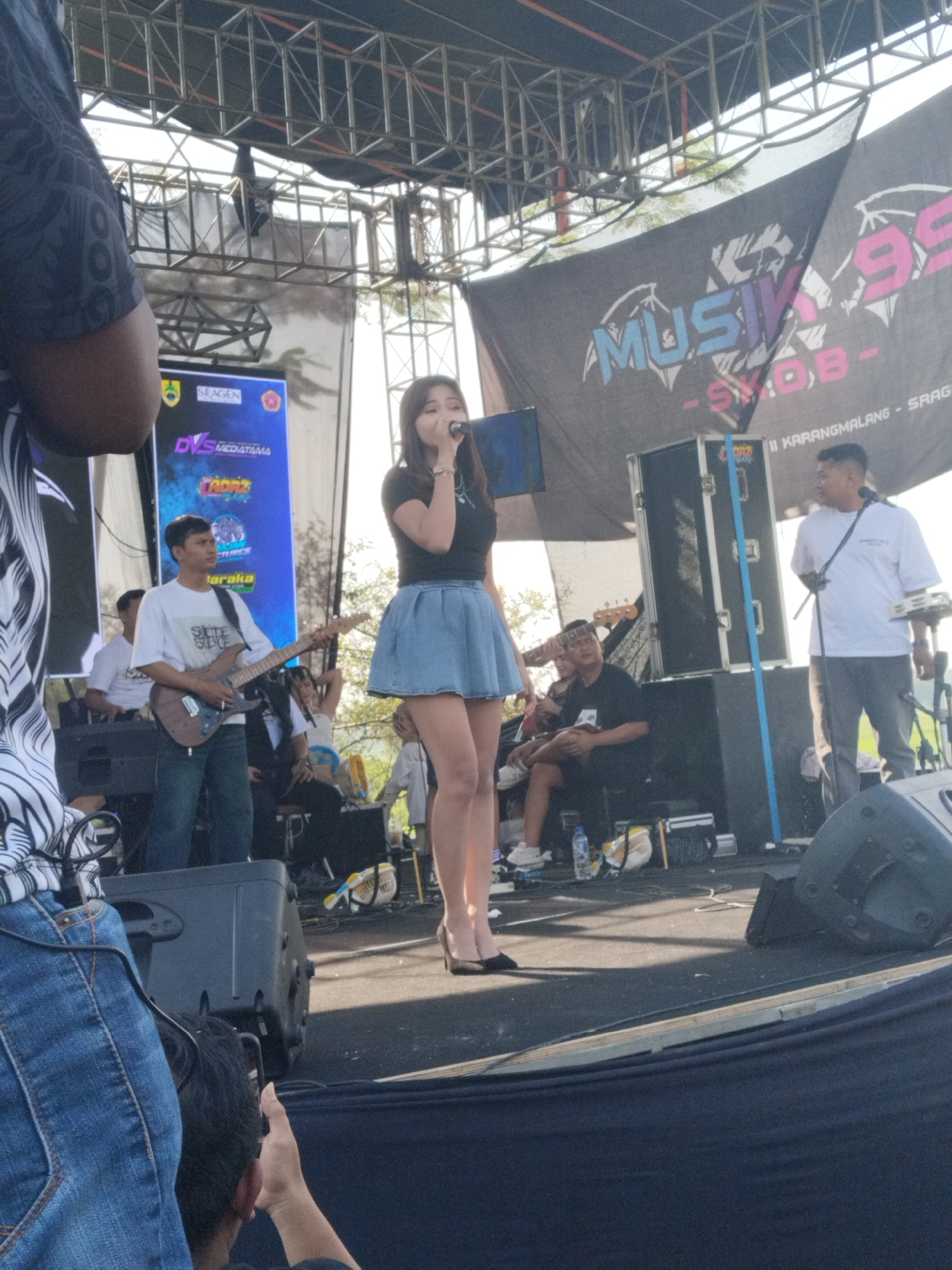 The image shows a live music performance taking place on an outdoor stage, likely in the area of Karangmalang-Sragen in Indonesia based on the banner in the background. A woman is singing into a microphone at center stage, dressed in a black top and a light blue denim skirt, with black high heels. Behind her, a male musician is playing an electric guitar. To the right of the singer, another man stands with what appears to be a keyboard and a microphone, and to their left are musicians and equipment. The stage has speakers and other music equipment. The background also has the "Musik 99 Syob" banner. The photo is taken from an audience member's perspective, with portions of two people in the foreground, one on the left side wearing a black-and-white patterned shirt and jeans, and another person's head visible near the bottom. The backdrop includes banners displaying the logos of the event organizers and sponsors like "DIS Mediatama" and "TRORZ". The stage is covered with a dark-colored material, and overhead is a steel framework supporting the stage lighting and equipment.