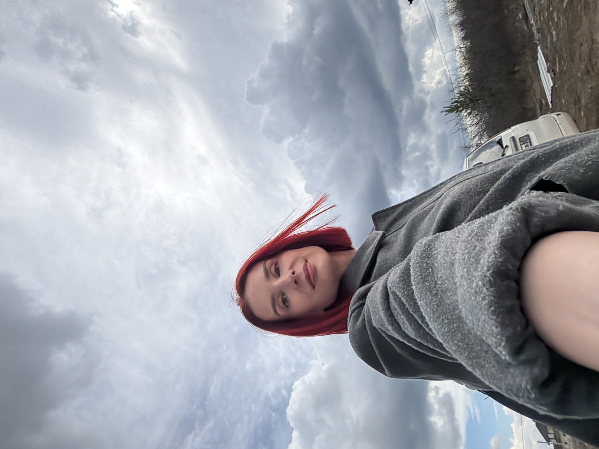 A young woman with short, bright red hair, slightly windswept, is featured prominently in this low-angle outdoor portrait. She looks upwards towards the camera with a calm, subtle smile. Her upper torso is visible, clad in a thick, textured grey long-sleeved garment, possibly a coat or sweater.
The scene is set outdoors under an expansive, heavily clouded sky. Large, grey, voluminous clouds dominate the sky, with occasional brighter patches suggesting an overcast daytime. In the background on the right side, a white utility van is partially visible on an unpaved or dirt surface. Behind the vehicle and extending across the horizon are numerous bare trees and bushes, indicating a cooler climate or season. Faint power lines can be observed against the sky above the tree line. The image captures a casual moment in what is likely Chaikovsky, Russia, with the woman appearing content despite the overcast weather. There is no visible text in the media file.