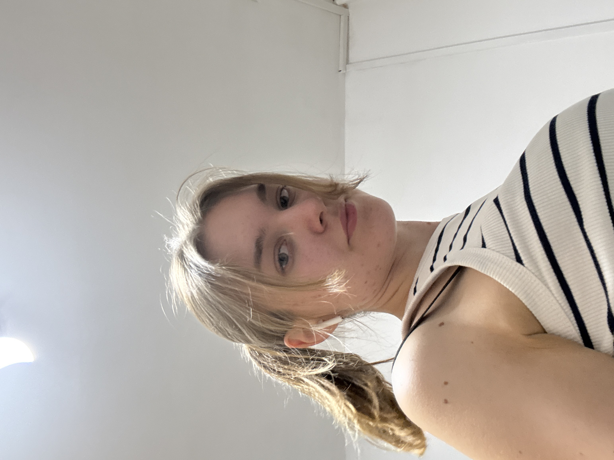 A woman with blonde hair is looking down at the camera. She is wearing a striped top. The background is a white wall. There is no indication of Perm, Russia in the image.