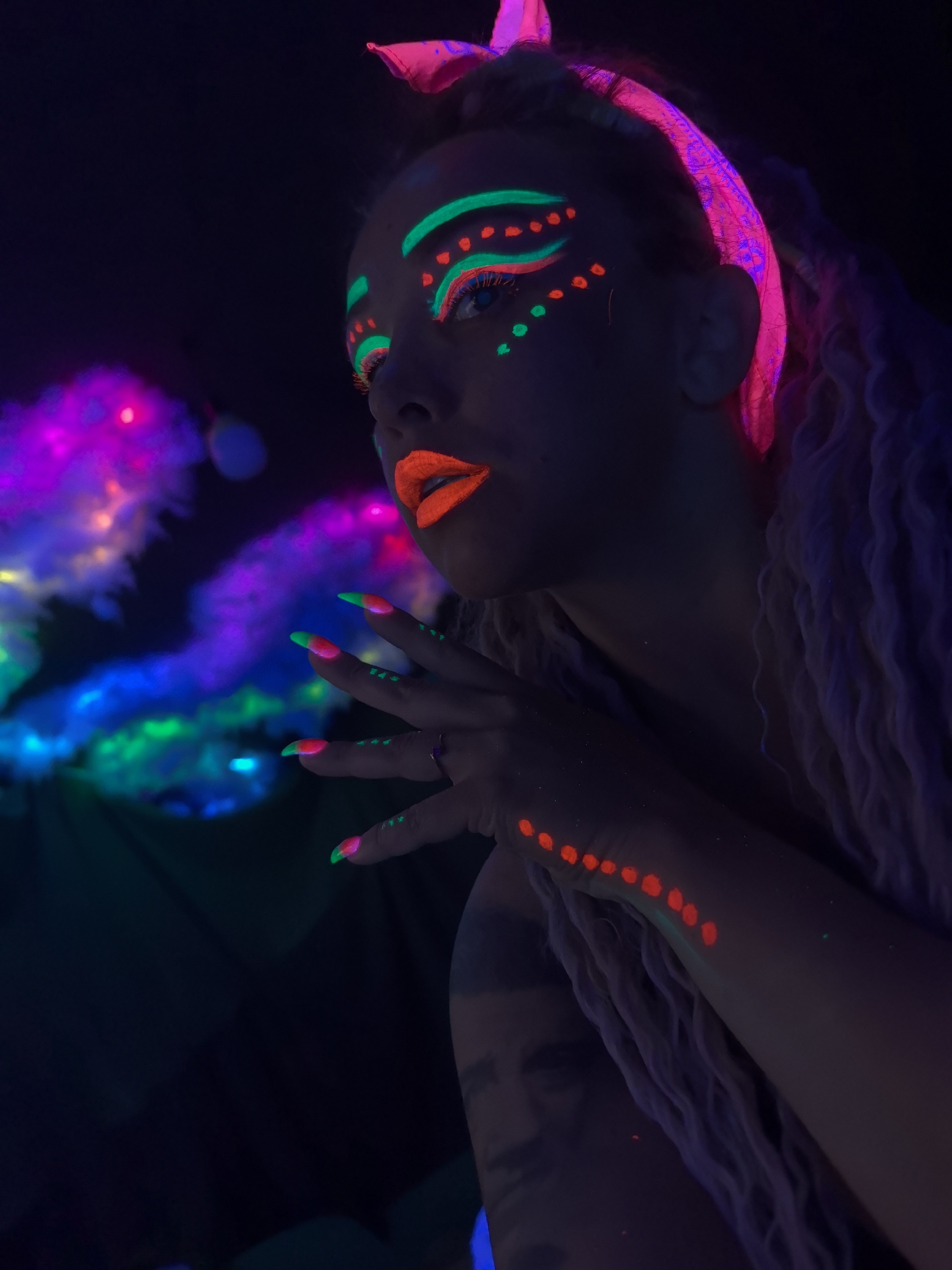 A low-angle close-up shot shows a fair-skinned person with fluorescent makeup in a darkened setting. The subject has neon green lines above and below the eyes, and small red dots between these lines. Their lips are painted bright orange, and their fingernails are neon green at the tips and red closer to the base. A row of red dots extends down the forearm. They wear a pink headscarf and have long, light-colored braids or dreadlocks. The person's hands are near their face, and they are looking off to the side. To the left, there is a blurry, multi-colored light display of purple, blue, and green. A tattoo is visible on their upper arm. The photograph features blacklight effects. This image does not provide information about the location in the city of Noviy, Russia.