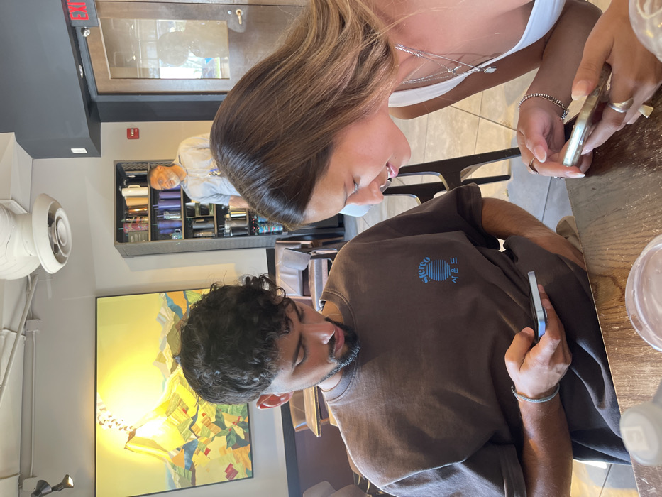 The image shows three individuals inside a commercial establishment, likely a cafe or coffee shop. In the foreground, a young adult male with dark curly hair and a short beard is seated at a wooden table, wearing a brown t-shirt with "Stereo" and Korean characters ("사운드") printed in blue. He is looking down at a light blue smartphone held in his right hand. To his right, a young adult female with long light brown hair is seated, wearing a white top and multiple silver necklaces. She is holding a light-colored smartphone and looking towards the male's general direction or his phone. Both foreground subjects are focused on their mobile devices. In the background, an older adult male with gray hair and a beard, wearing a light blue collared shirt, stands behind a dark shelving unit filled with various mugs and tumblers. He is looking to the right. On the left background wall, a large, abstract painting depicting a yellow sun-like form over a colorful geometric landscape is visible. A white ceiling fan with a light is mounted overhead. A wooden door with a glass panel and a partial "EXIT" sign is visible on the far right. The specific location within the city of Washington, United States, cannot be determined from the image content.