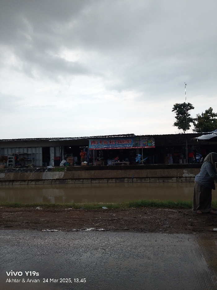 The image shows a view of a street in Kecamatan Haurgeulis, Indonesia (latitude: -6.419, longitude: 107.972). There are multiple shops or stalls lining the street. A sign above the shops reads "Baju & Tas Perempuan", which means "Women's Clothes & Bags" in Indonesian. The street is wet and muddy with a canal running along the side. A few people can be seen walking in the distance. The sky is overcast with grey clouds. The time is 13:45.