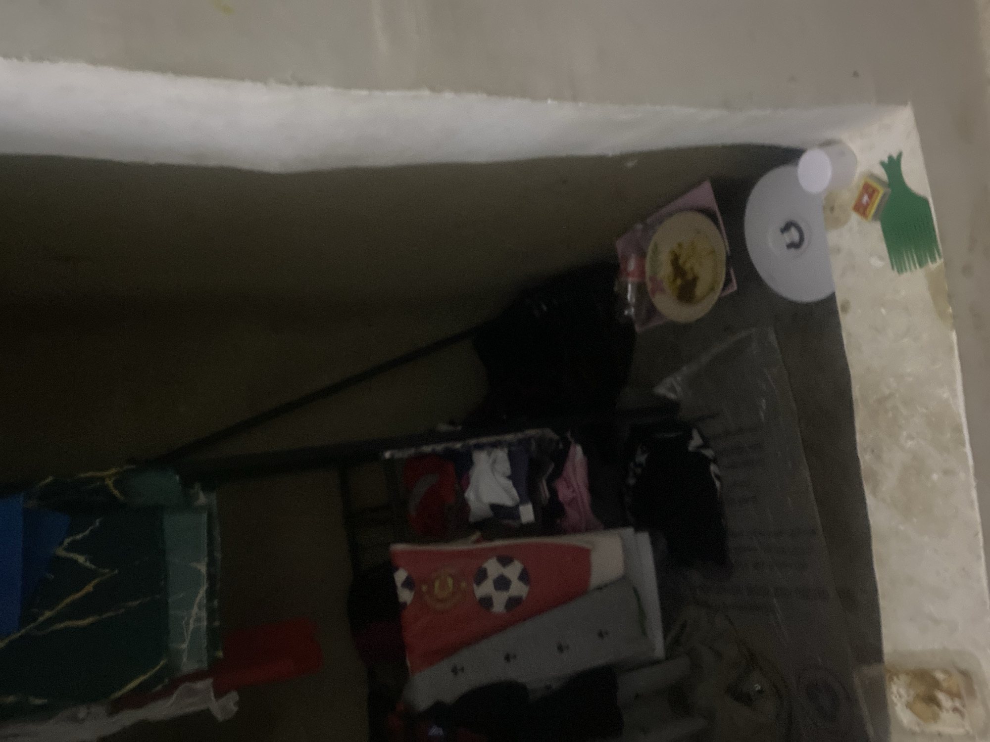 The image depicts a dimly lit interior space, possibly a room or storage area, in Howayi, Nigeria. The scene is cluttered with various items, suggesting a state of disarray or storage. In the foreground, a beige-colored surface is visible, with a small white and brown object on it, potentially a food item. To the left, a white, circular object with a dark "U" shape is present, possibly a lid or base. Next to it, a small white cup is standing upright. A green object resembling a comb lies on the surface. A small box with a yellow and red design is also visible. In the background, a dark backpack is placed on the floor. Various pieces of clothing are scattered around, including a red garment with white soccer ball designs, a gray garment with black patterns, and other folded clothes. A metal shelf unit is partially visible, supporting some of these items. The walls appear to be a light, textured material. The overall impression is one of a utilitarian and somewhat neglected space.