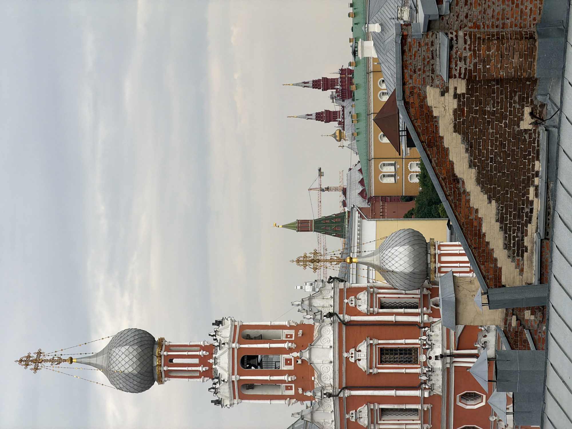 This is a medium-shot, eye-level, exterior, vertical photograph of a cityscape in Moscow, Russia. The image captures a cluster of architectural structures, featuring intricate details characteristic of Russian Orthodox churches and historical buildings. In the foreground, a red building with white decorative elements and a silver onion dome dominates the left side of the frame. This structure appears to be a bell tower, with visible bells inside its open belfry. Adjacent to it, a smaller silver onion dome with a golden cross is visible.
In the middle ground, the red building gives way to a complex of other buildings. A green-roofed structure is prominent, and further in the distance, several towers with distinctive red brickwork and spire-like tops are visible, one of which has a golden cross. Two construction cranes are also present, suggesting ongoing development or restoration work in the area. The background is filled with a cloudy, overcast sky. The overall atmosphere of the image is one of historical depth and architectural grandeur, typical of central Moscow. No people are present in the image.