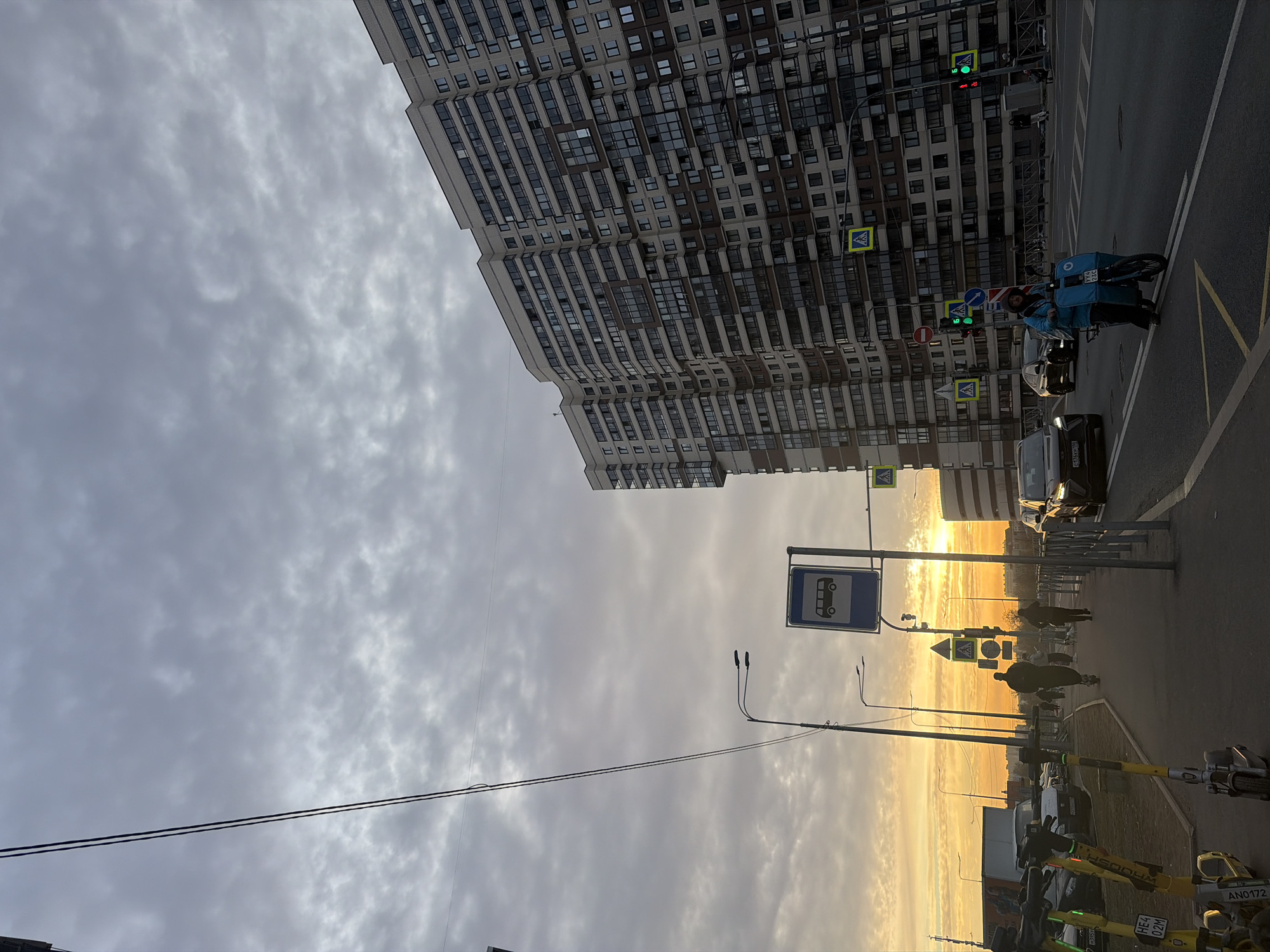 The image shows a street scene in Saint Petersburg, Russia. A tall, modern apartment building occupies the upper portion of the frame. Utility poles and wires stretch across a sky filled with overcast clouds. A bus stop sign is visible. The horizon is illuminated by a sunset. A delivery worker on a bicycle is waiting at a crosswalk. Motor vehicles are on the road, and pedestrians are walking along the sidewalk. Parked scooters are visible in the foreground.