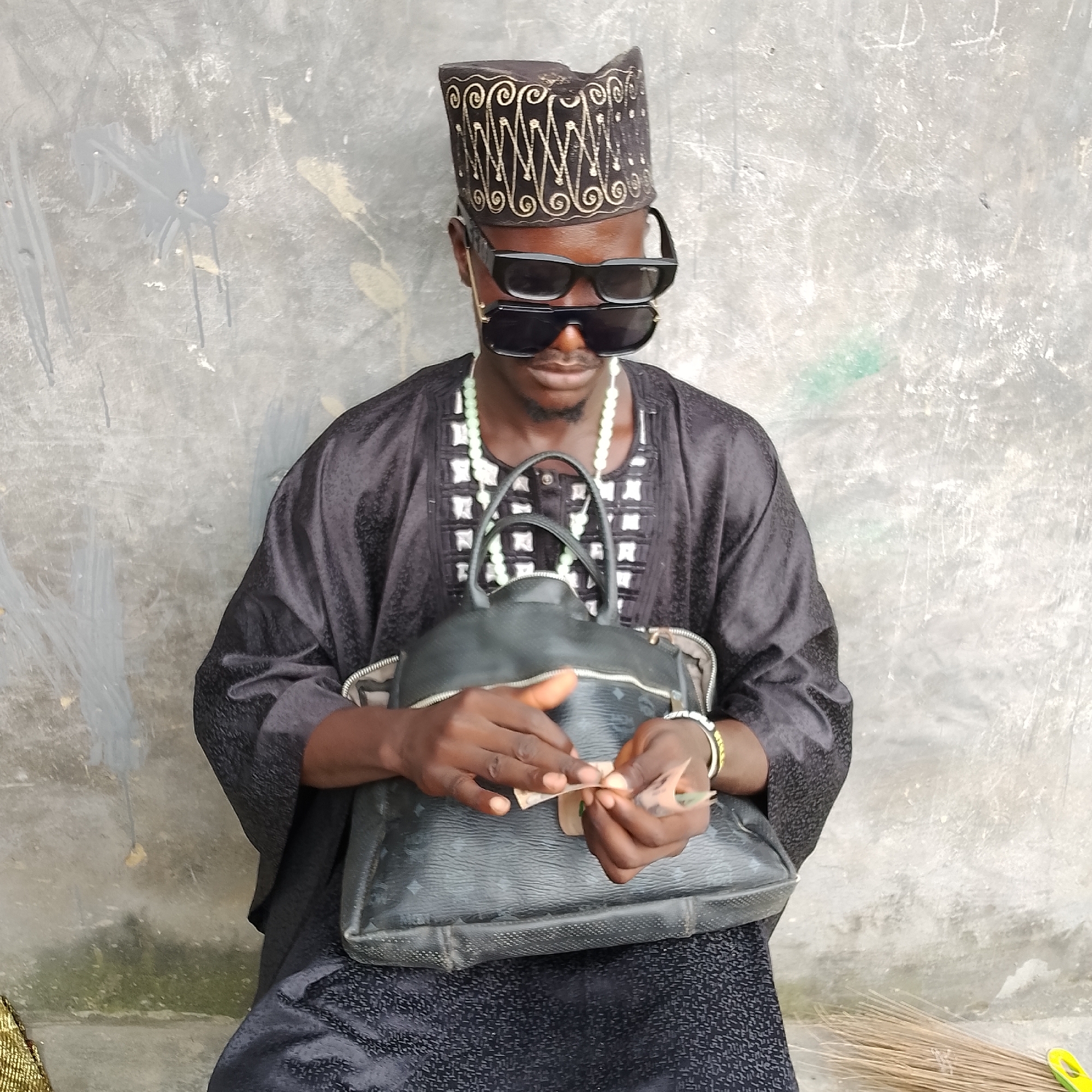 A man, seated in front of a grey, textured wall, wears a black, ornate cap, two pairs of sunglasses stacked on his face, and a black traditional garment adorned with white geometric patterns. He is holding cash in his hands, appearing to be counting or organizing it, over a black leather bag resting on his lap. The environment suggests an outdoor setting in Ado, Nigeria, with a broom visible on the right side of the frame, leaning against the wall.
