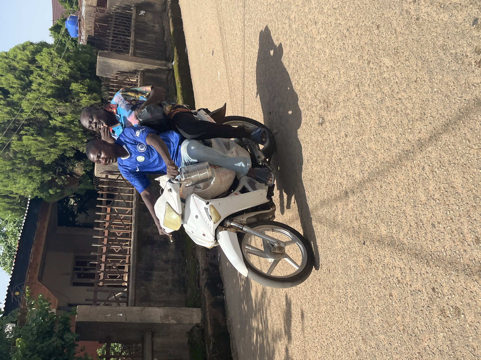 Two young men are riding on a white motorbike on a paved road in Jos, Nigeria. The man in front is driving and is wearing a blue t-shirt with the Chelsea FC logo. He has a metal tiffin carrier attached to the side of the motorbike. The man behind him is wearing a colorful, patterned shirt and appears to be laughing. The background shows houses with corrugated iron roofs and a fence. Trees and foliage are visible, suggesting a residential area. The day is bright, with shadows cast on the road.