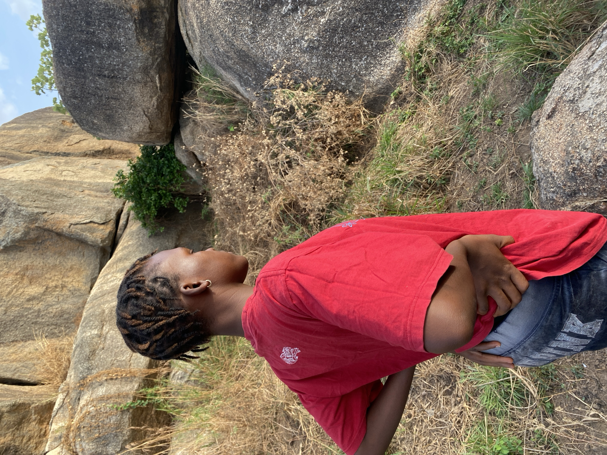 A young woman stands outside in a rocky, grassy area. She has braided hair, wears a small hoop earring, a red t-shirt, and dark jeans with what appear to be rhinestone embellishments. Her arms are crossed. The background includes large rocks and vegetation. The setting is likely near Vom, Nigeria, based on the visual characteristics of the landscape.