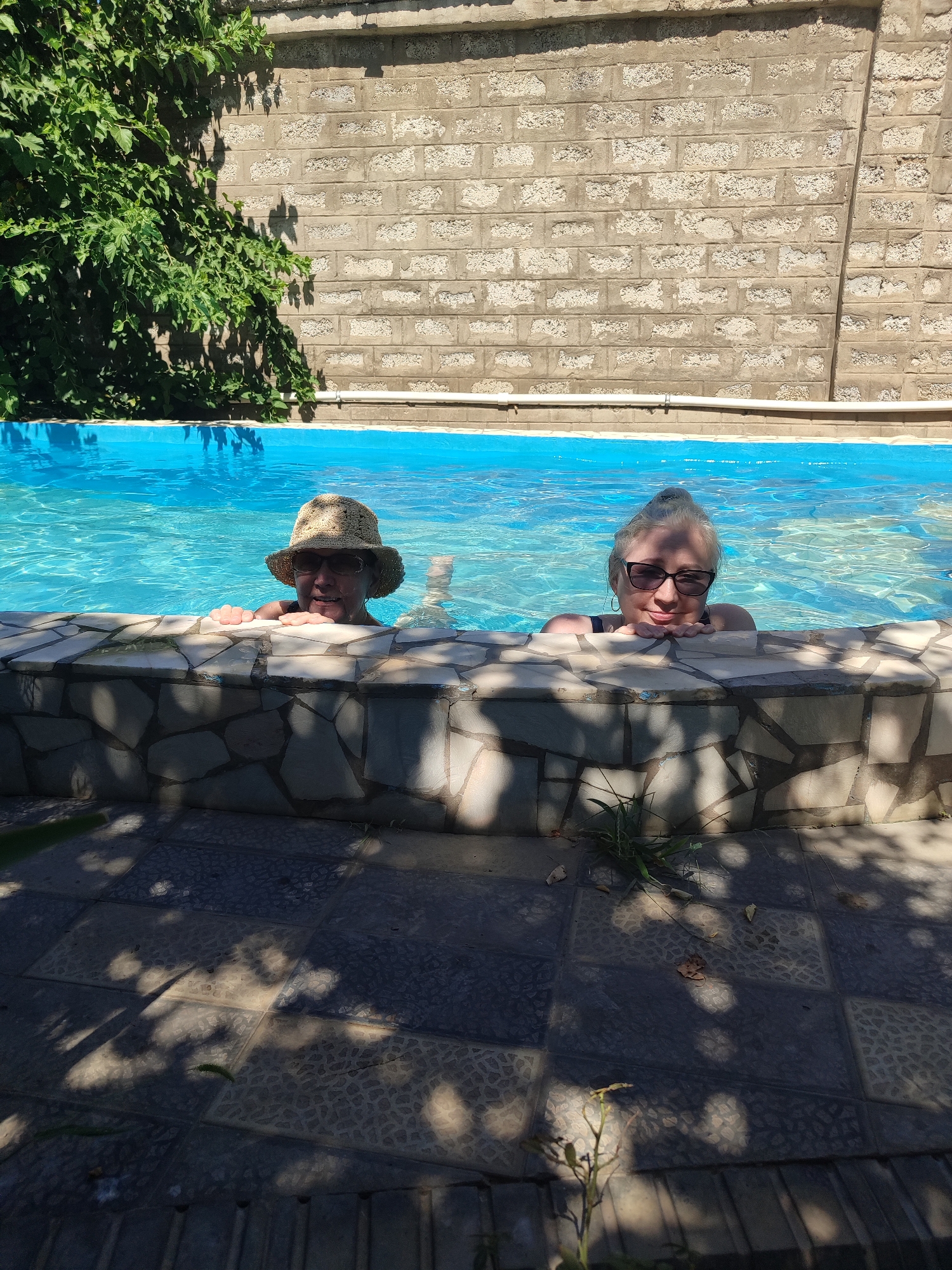 Two women are pictured in a swimming pool, leaning on the stone border of the pool. The woman on the left is wearing a straw hat and sunglasses. The woman on the right is wearing sunglasses. The pool is filled with blue water and reflects sunlight. Behind the pool is a wall constructed of stone blocks, with a green vine growing on the left side. The ground in front of the pool is paved with dark tiles, and there are shadows cast by leaves on the ground. The scene appears to be outdoors on a sunny day. The image was taken in the city of Eshmataul, Uzbekistan.