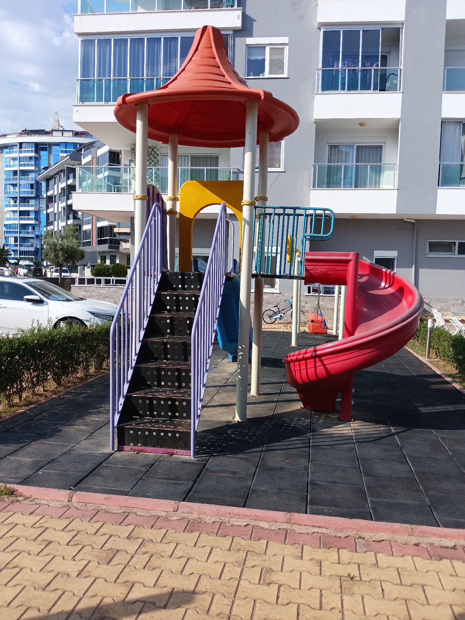 The image depicts an outdoor playground with a red spiral slide, a purple staircase, and a yellow slide, all part of a larger play structure. The playground is situated on a black rubberized surface, with a border of interlocking beige and pink pavers in the foreground. Behind the playground, a modern grey building with balconies and large windows rises. To the left, a blue building with many windows and a distinctive curved design is visible, along with parked white cars and a dense green hedge. The sky is blue with some white clouds, suggesting a clear, sunny day. The lighting indicates it's daytime, likely late morning or early afternoon. There are no people visible in the image, and no text is present. The scene appears to be a residential area, possibly an apartment complex, in Alanya, Turkey, given the architectural styles and the presence of a playground.
