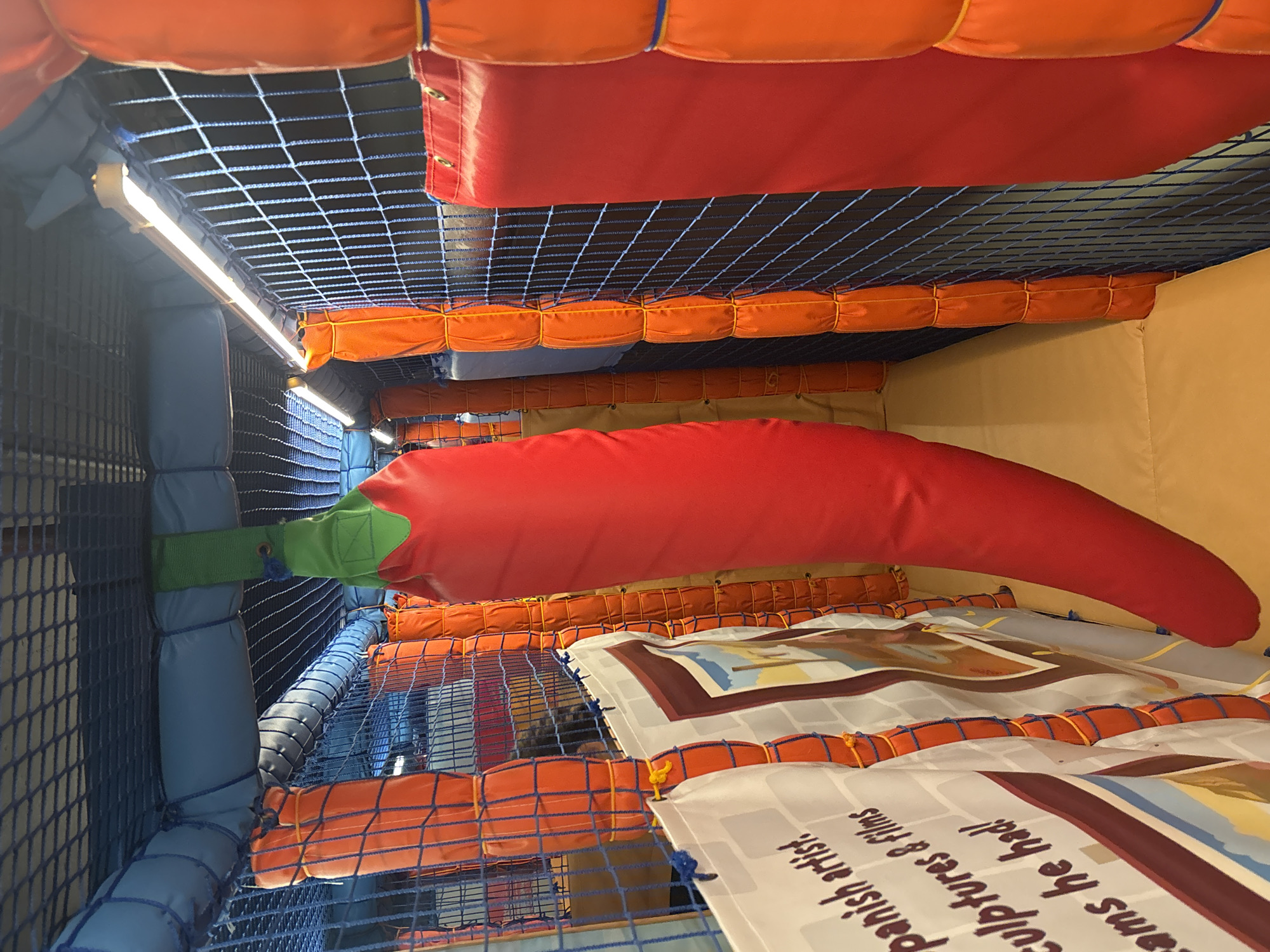 The image shows a section of a soft play area, likely a children's indoor playground. A large, inflatable chili pepper decoration hangs from the ceiling in the center of the frame. The area is enclosed by netting and padded orange and red barriers. Fluorescent lights are visible along the ceiling. On the left, a banner with text and images is partially visible. The floor is covered with a yellow padded mat. The scene appears to be indoors, with no external environmental elements visible. There are no people or animals depicted in the image. The location is not specified as Dubai, United Arab Emirates.