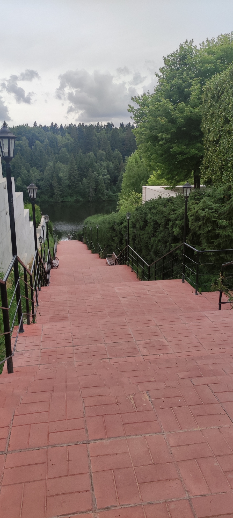 A wide, downward-sloping staircase paved with red bricks leads toward a calm body of water. The staircase is bordered by black metal railings on both sides, and several black, old-fashioned lampposts are visible along the railing. Benches are placed on the sides of the stairs. Dense green trees line the far bank of the water, creating a natural backdrop. The sky is overcast and gray. A single person is visible at the bottom of the stairs, near the water's edge. The overall scene suggests an outdoor park or recreational area.