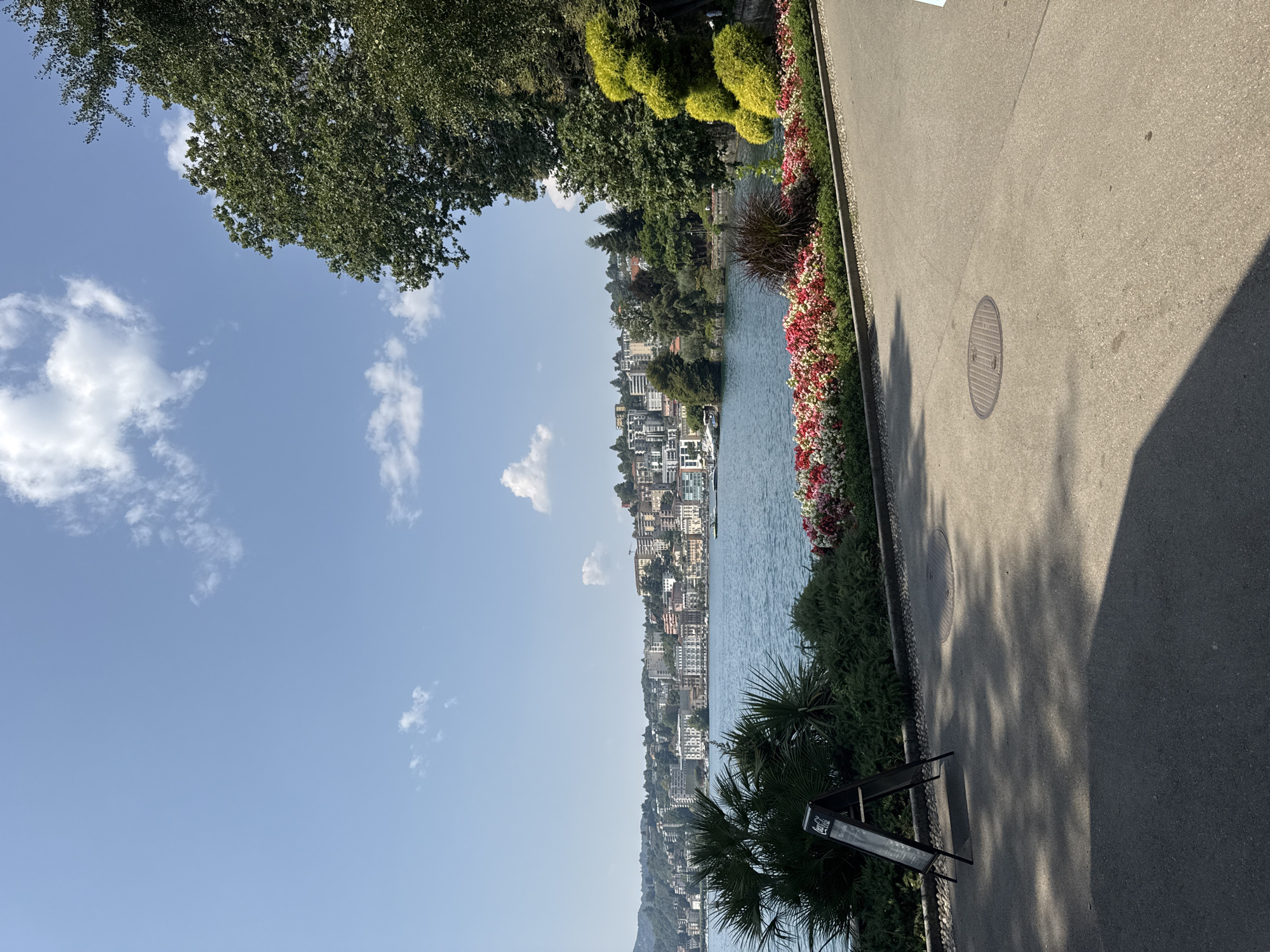 The image depicts a waterfront view of Lugano, Switzerland, on a clear day with scattered clouds. In the foreground, a paved walkway with a manhole cover is visible, partially shaded by trees. To the left, a black A-frame sign with a Coca-Cola logo stands near lush green foliage and palm trees. A vibrant flower bed with red, pink, and white flowers runs along the edge of the walkway. Beyond the flowers, a calm blue lake stretches out, with a cityscape of buildings, some with light-colored facades and balconies, lining the opposite shore. The buildings are situated on a hillside that extends upwards, covered in trees and residential structures. The sky is a bright blue with white clouds.