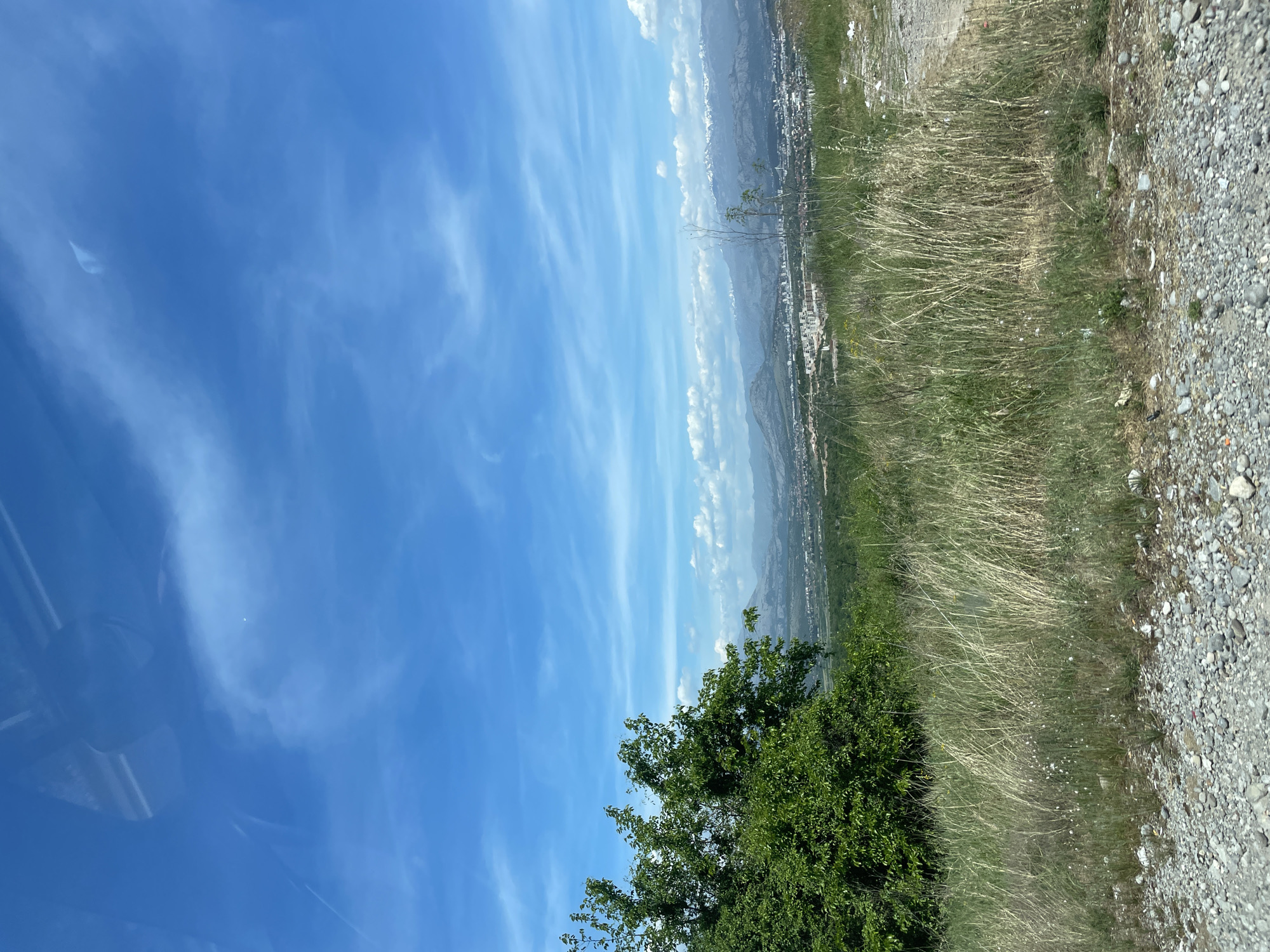 The image, oriented horizontally, displays a sweeping landscape view captured on a clear, sunny day. A vast expanse of bright blue sky dominates the upper portion, adorned with delicate, wispy white clouds that stretch across the frame.
In the foreground, a vibrant green, leafy tree or large bush is visible on the left side. To the right, the ground transitions from patches of dry, tall grass and some greener foliage to a rough, stony, and gravel path or shoulder along the far right edge.
The midground reveals an expansive valley or plain dotted with numerous buildings, indicating a town or city nestled in the distance. Beyond this urban sprawl, the background features imposing mountains, some appearing rugged and possibly snow-capped in places, rising under a clear sky.
No people or specific activities are visible, presenting a static, natural scene. The bright light suggests it is daytime. The vantage point overlooks a broad geographical area, consistent with a view from an elevated position such as those found around Gornji Kokoti, Montenegro, offering a panorama of the region's urban and mountainous terrain. No text is visible within the image.