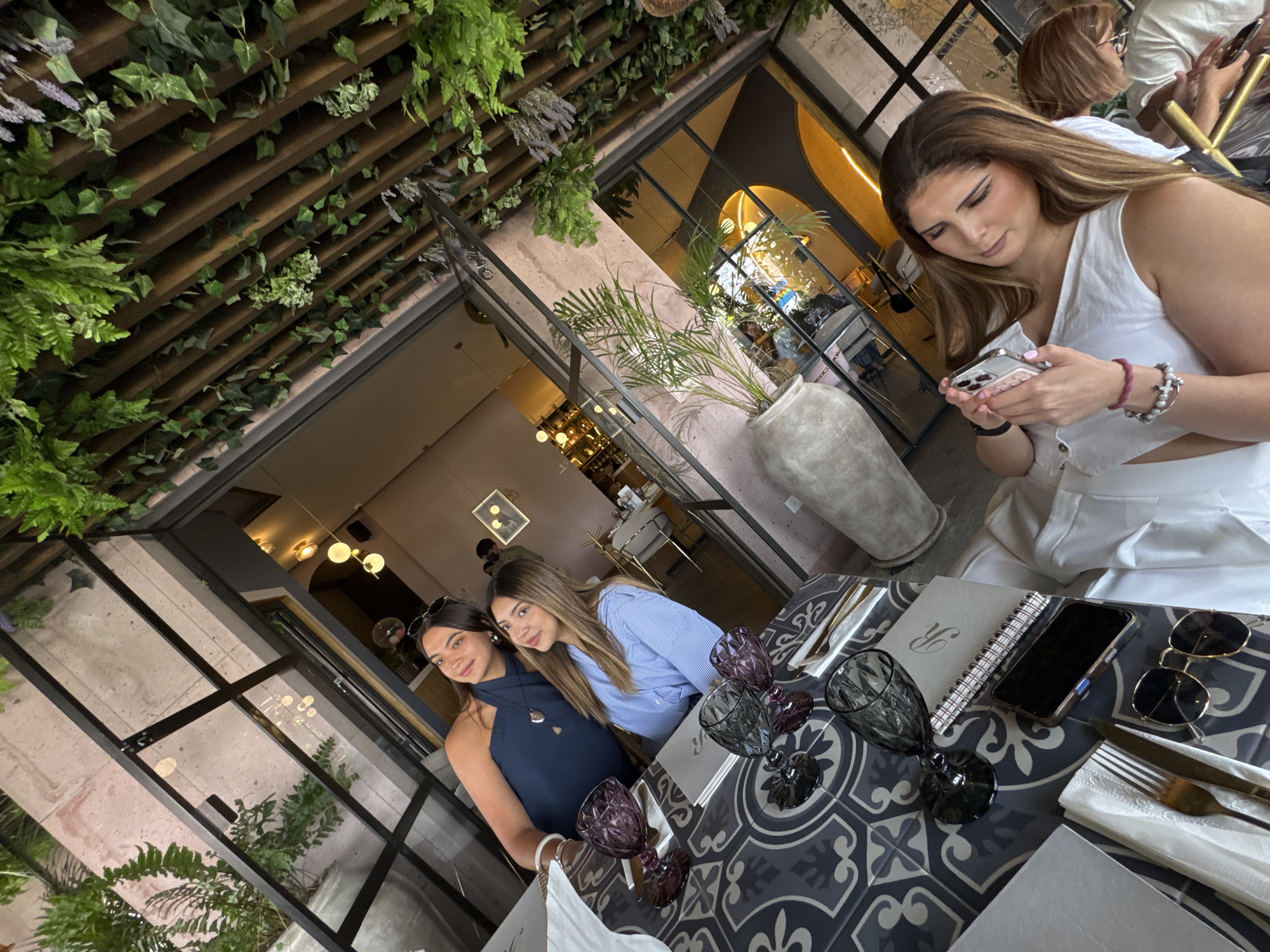 This media file captures a social gathering of three women in a stylish restaurant or cafe setting, likely in Puebla, Mexico. The overall scene suggests a blend of indoor comfort with a semi-outdoor, garden-like ambiance.
In the foreground to the right, a woman with long brown hair, wearing a white sleeveless top, is intently looking at her smartphone. She has multiple bracelets on her wrist, including beaded ones in white and purple tones. Her attention is solely on her device.
Towards the mid-ground on the left, two other women are seated at a table, both looking towards the camera with pleasant expressions. One woman, with dark hair and sunglasses perched on her head, wears a dark blue halter-neck top. Next to her, another woman with long brown hair wears a light blue striped collared shirt.
The setting is characterized by its lush greenery and modern architectural elements. Above, a ceiling features a wooden slatted structure adorned with various artificial or real plants, including ferns, ivy, and sprigs resembling lavender. Large glass panels and dark window frames divide the space, revealing glimpses of the interior and possibly an exterior area. Potted plants, like a palm in a large textured pot, further enhance the green aesthetic.
The table in the foreground features a distinctive black and white patterned tile surface. It is set with ornate purple and dark grey/black stemmed glasses, silver cutlery, and white napkins. A smartphone, a pair of sunglasses with dark lenses, and a small spiral-bound notebook are also on the table. The notebook has a stylized "R" visible on its cover. In the background, modern light fixtures, including globe pendants and arched doorways, contribute to the contemporary design. Another individual is partially visible in the far background.
The lighting appears to be natural mixed with ambient interior lights, suggesting it could be daytime, possibly late afternoon or early evening. The overall atmosphere is relaxed and social.