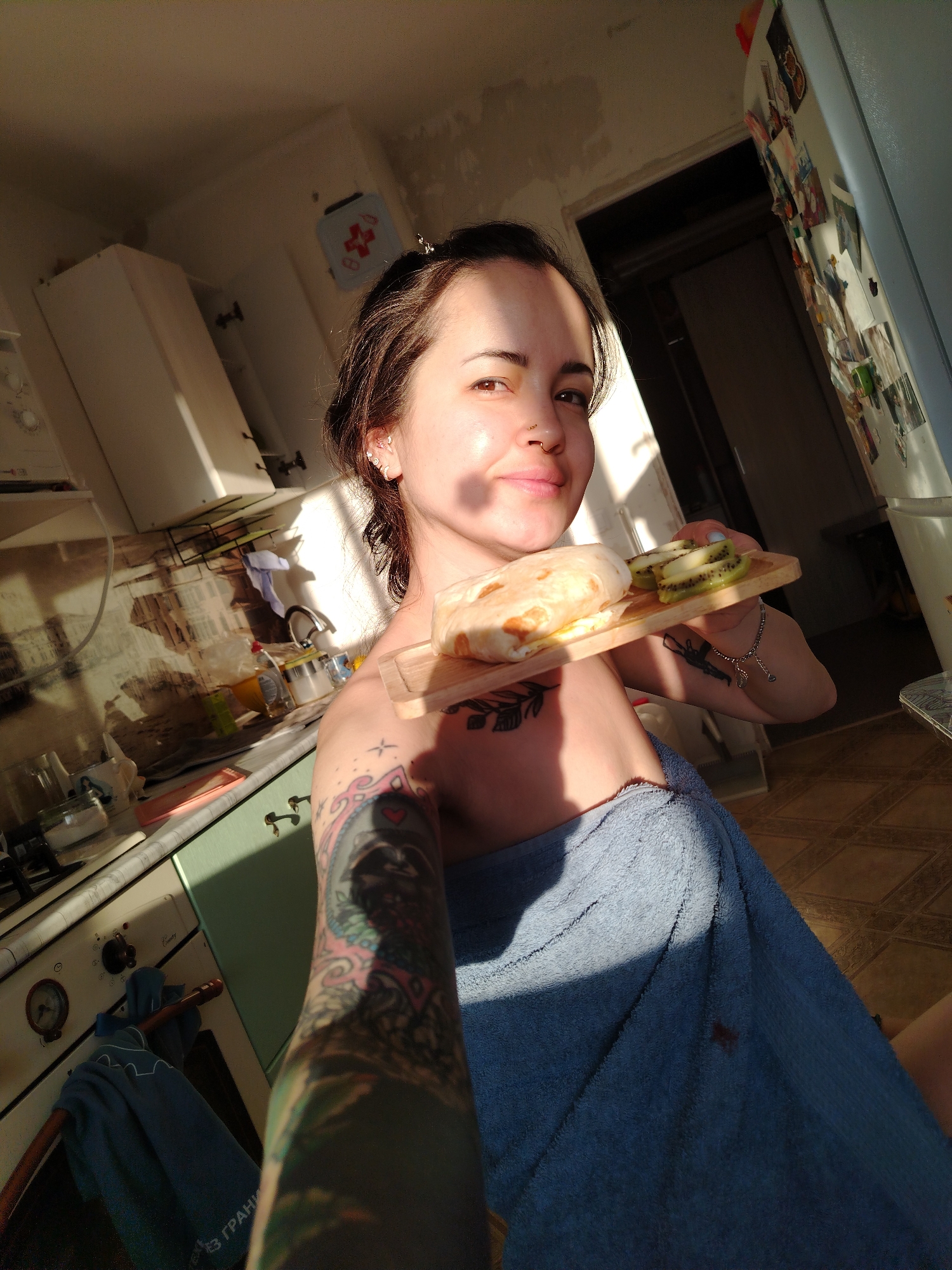 A person with brown hair and multiple ear piercings is holding a wooden board with a wrapped burrito and sliced kiwi on it. The person is wearing a blue towel wrapped around their body and has multiple tattoos on their arms and chest. The setting appears to be a kitchen, featuring a stove, cabinets, a refrigerator with magnets, and tiled floors. The scene is illuminated by bright sunlight. The location is Unknown, Unknown.