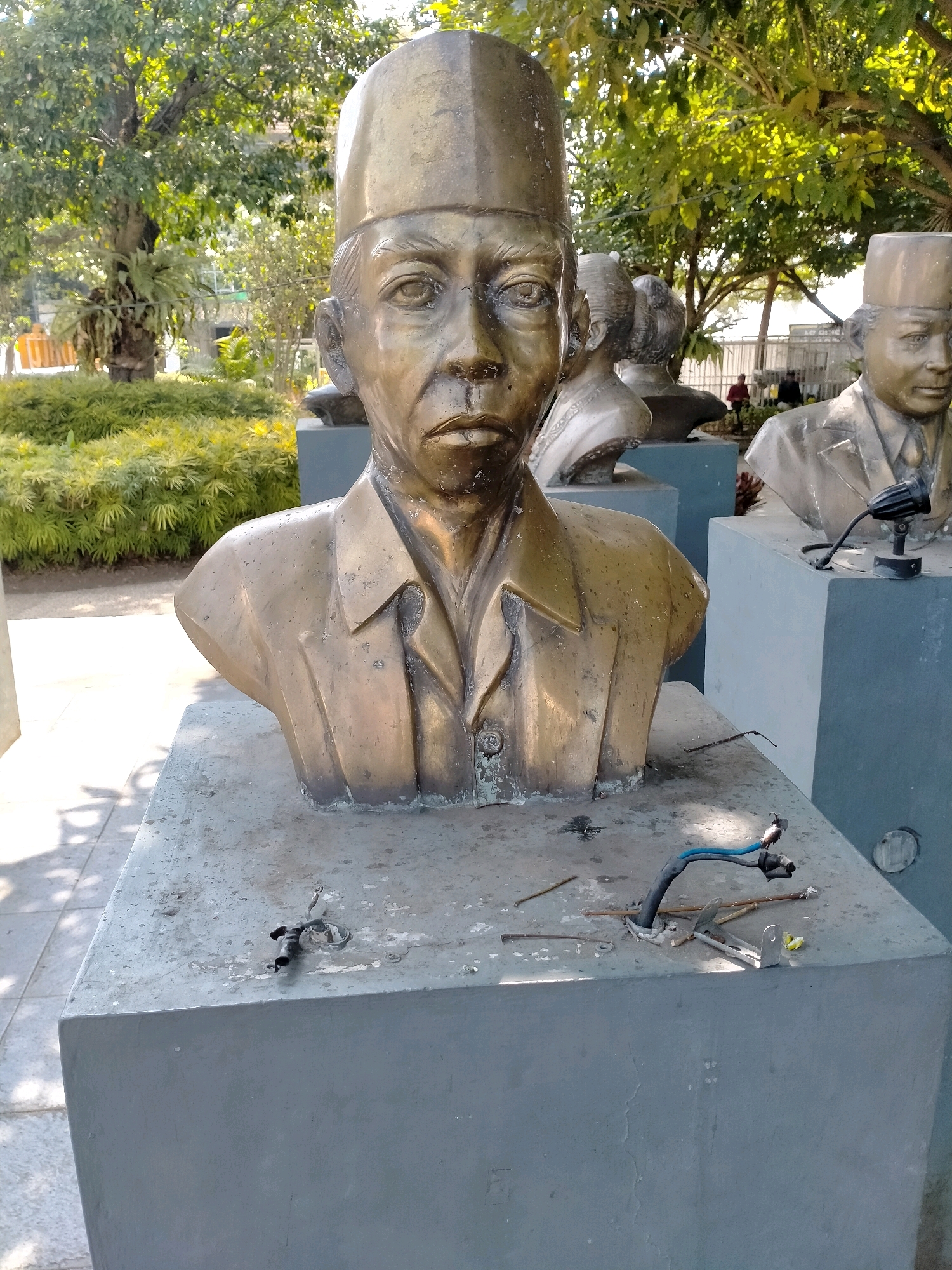 A bronze bust of a man wearing a cap stands prominently in the foreground, positioned on a grey concrete pedestal. The bust is detailed, showing the man's facial features and attire, which includes a collared shirt and a buttoned jacket. Behind this central bust, other bronze busts are visible, suggesting a monument or a collection of historical figures. The busts are situated outdoors, with lush green vegetation and trees in the background, indicating a park or garden setting. Sunlight casts shadows on the ground, illuminating the scene and highlighting the metallic sheen of the sculptures. The concrete pedestal of the main bust is littered with debris, including wires and small metal objects, suggesting possible damage or disrepair. The location is identified as the "Taman Rekreasi Selecta" in the city of Batu, Indonesia.