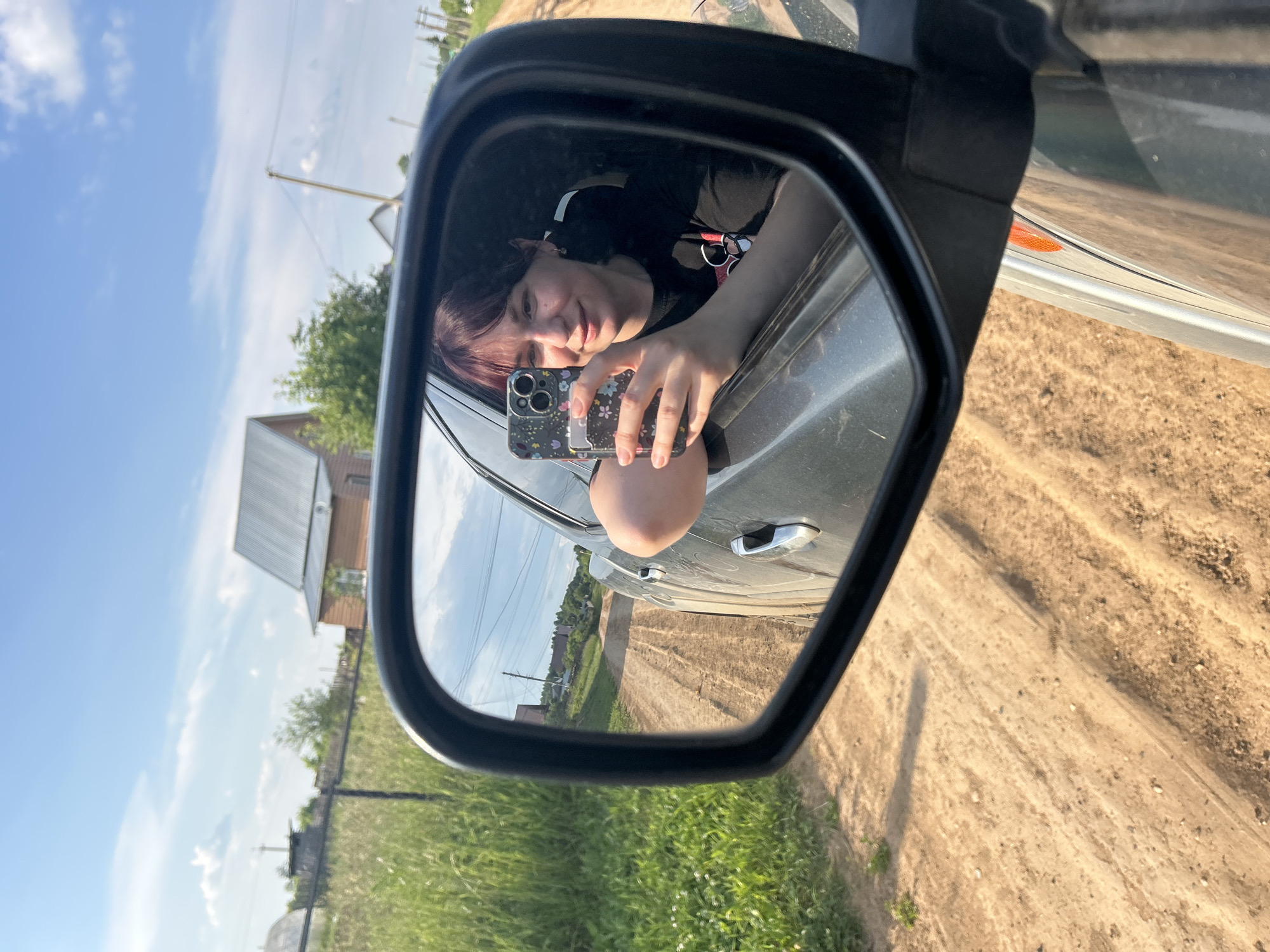 The image is a portrait-oriented selfie taken from inside a car, reflected in the side-view mirror. The main subject is a young person with dark hair, possibly dyed with purple streaks, leaning out of the car window. They are holding a smartphone, also visible in the reflection, and taking the photo. The person is looking at the camera with a relaxed, slightly smiling expression. They are wearing a dark t-shirt with a graphic design.
The background reflected in the mirror shows a rural setting. There is a dirt road with tire tracks, leading away from the car. On either side of the road, there is green grass and vegetation. In the distance, there are houses and trees. The sky is blue with scattered white clouds, suggesting it is daytime and the weather is fair. The presence of a dirt road and the type of buildings visible are consistent with a rural location in Russia, specifically Фоки. The reflection also shows the side of the car, including a door handle and an orange indicator light. The image captures a moment of leisure or travel.