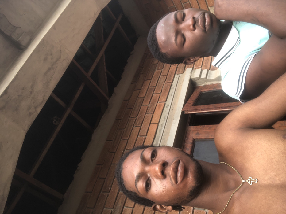 Two dark-skinned men are positioned in the frame. One wears a light-colored, collared shirt with white stripes, eyes closed. The second is shirtless, wearing a gold chain with a cross pendant. They appear to be outside. A brick building and visible ceiling joists form the backdrop. The camera angle is low, tilted upward.