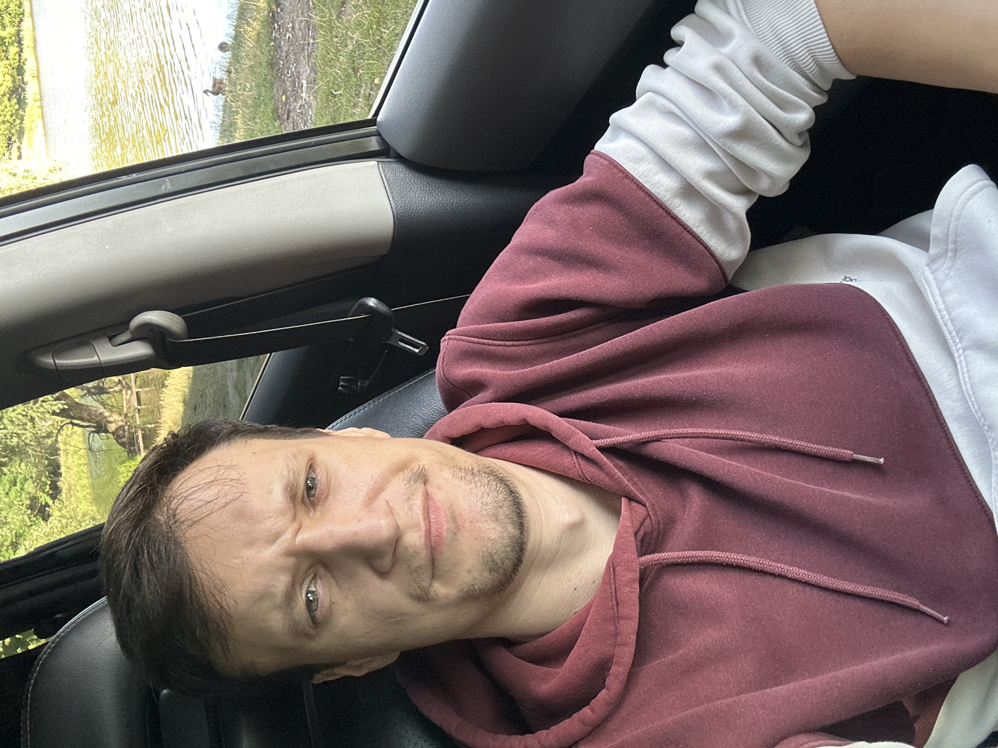 A man is seated in the driver's side of a vehicle. He is wearing a maroon and white hooded sweatshirt. The interior of the car is visible, including the black leather seat and the gray pillar. Outside the car window, a body of water with ripples is seen, with trees and grassy banks along the edge. Two ducks are visible on the grassy bank near the water's edge. The location is identified as Makiv, Ukraine.