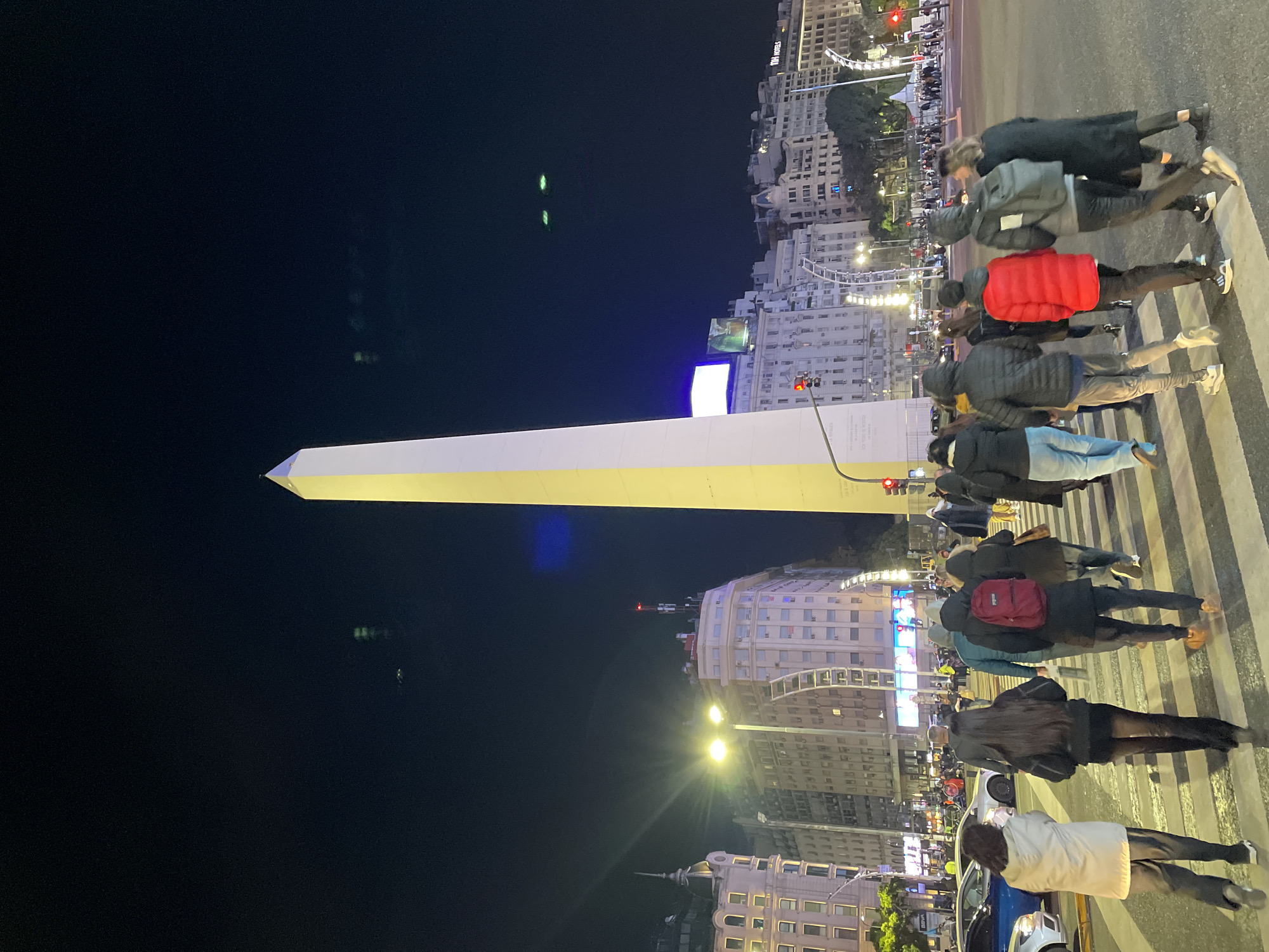 The image depicts the Obelisco, a historical monument in Buenos Aires, Argentina, at night. The Obelisco is lit up and stands prominently against a dark sky. Several people are crossing the street in front of the monument, walking on a zebra crossing. Buildings with lighted windows line the streets in the background. There are streetlights and traffic signals visible. A few cars are parked near the crosswalk.
