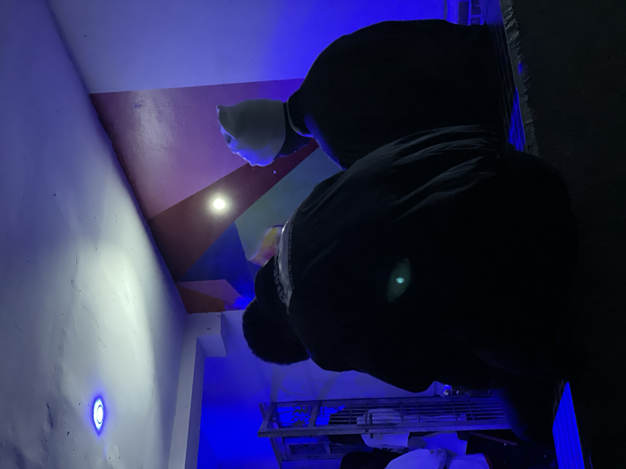 The image depicts an indoor scene with low lighting, dominated by blue and purple hues. Two individuals are seated, viewed from behind. The person on the right wears a light-colored beanie and a dark jacket. The person on the left wears a dark jacket with a visible white emblem on the back. In the background, a metal gate or fence with intricate designs is visible, illuminated by blue light. The wall behind the individuals features abstract geometric shapes in shades of red, purple, and blue. A circular light fixture with a bright blue glow is present on the ceiling. The overall environment suggests an urban setting within the city of Durimi, Nigeria, potentially a public or private gathering space, illuminated by artificial lighting.