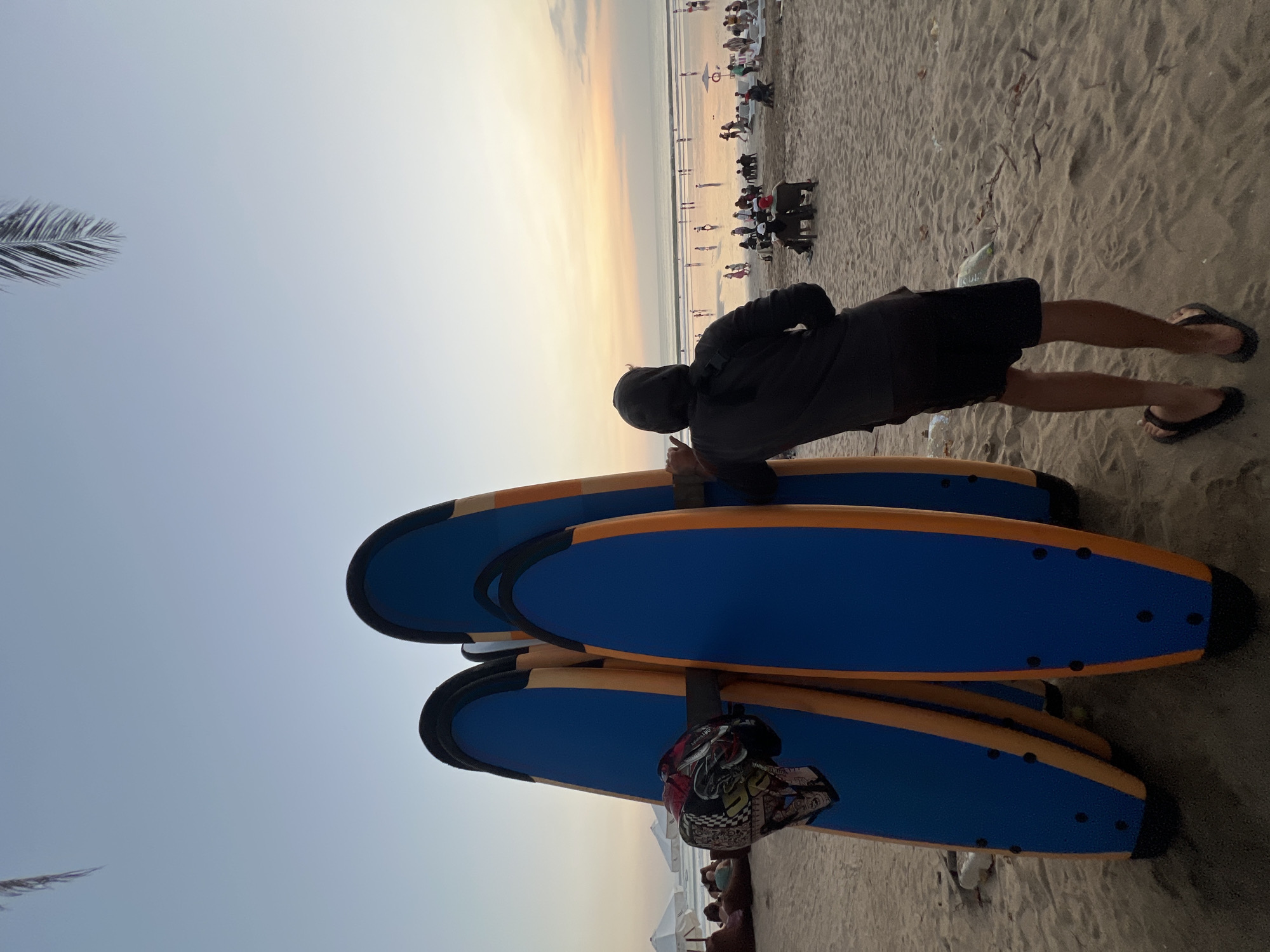 In Kuta, Indonesia, a person wearing a black hoodie and black shorts leans against a row of surfboards on a sandy beach. The surfboards, with blue tops and orange sides, are arranged upright. A helmet with a colorful, patterned design is attached to one of the surfboards. In the background, the ocean meets a pastel-colored sky at sunset. Numerous people are scattered across the beach and in the shallow water, some sitting on chairs or beanbags, while others are walking or standing. Umbrellas are visible in the distance. The scene captures a tranquil moment on a popular Indonesian beach.