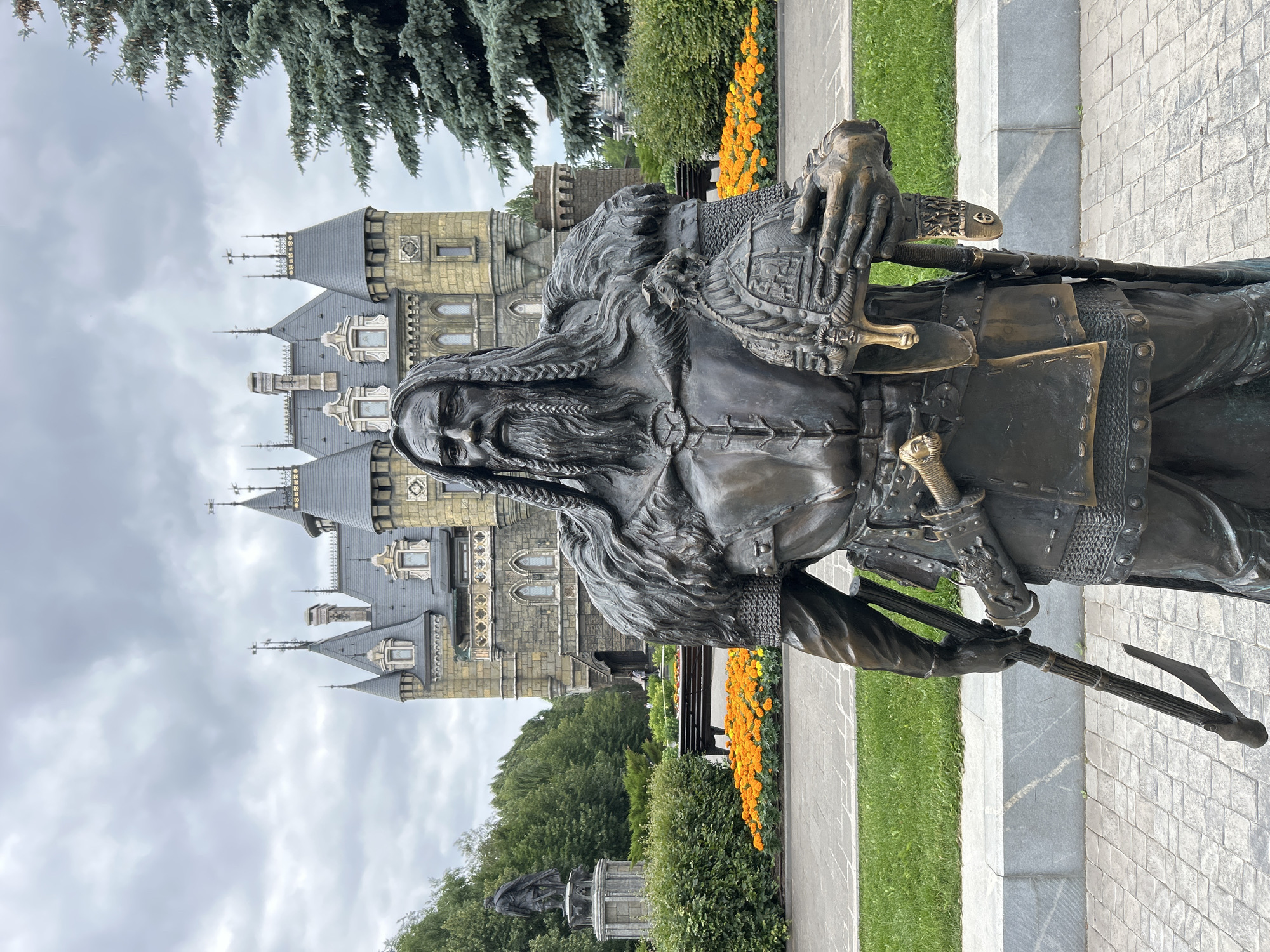 The image depicts a large bronze statue of an ancient warrior in front of a gothic-style building. The warrior is depicted with a long braided beard and hair, wearing armor and fur. He holds a helmet in his right hand and an axe-like weapon in his left. The building behind him appears to be a castle or a themed structure, with turrets and multiple windows. In the background, there are trees and a cloudy sky. The area in front of the building and statue is landscaped with grass, a paved walkway, and a flower bed of orange flowers. To the left of the statue, a smaller statue of a figure on a pedestal can be seen. This scene is located in the city of Khryashchevka, Russia, likely within the "Fairytale Town" amusement park, as the building resembles the park's central attraction. There are no visible people interacting with the statue or the building in the immediate foreground.
