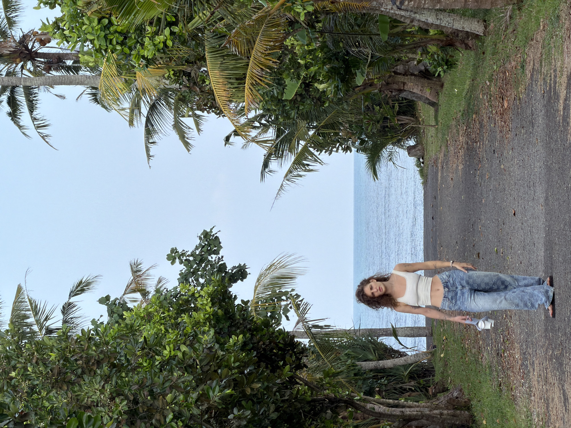 A young woman with curly brown hair stands on a paved path that leads towards the ocean. She is wearing a white crop top and loose-fitting blue jeans. She holds a white object in her left hand. The path is flanked by lush green trees and palm trees. The ocean is visible in the background under a pale blue sky. The image appears to have been taken in Kediri, Indonesia, given the tropical vegetation and coastal setting.