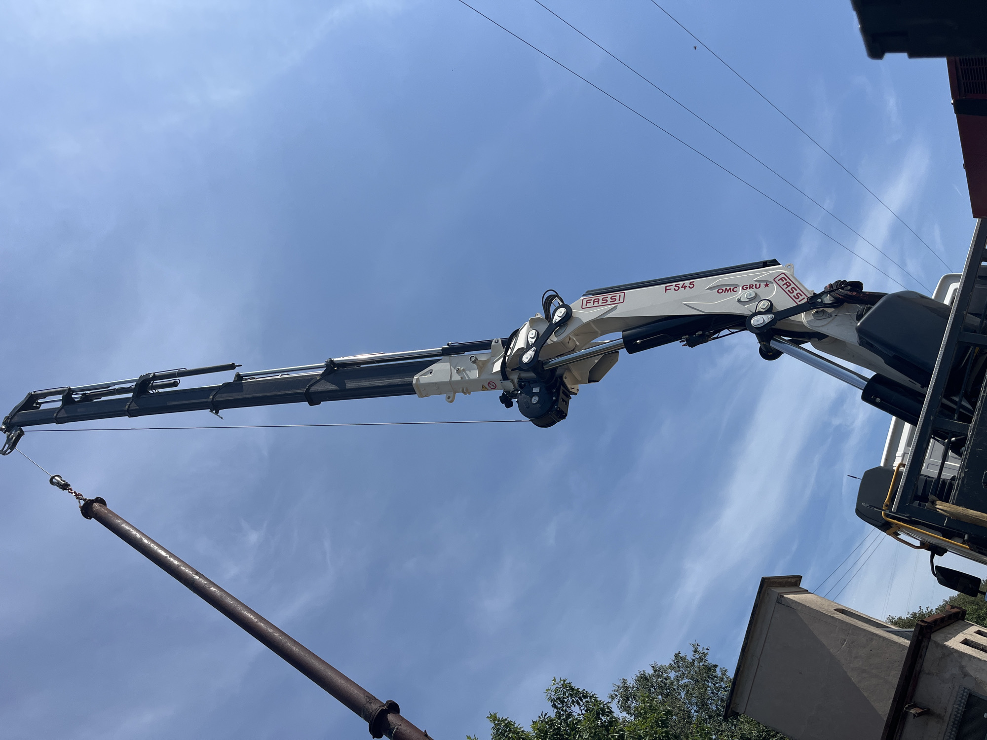 In Castiglione delle Stiviere, Italy, a large industrial pipe, rusted and weathered, is being lifted into the air by a white and grey Fassi F545 OMC GRU crane mounted on a truck. The crane's hydraulic arm is extended upwards, suspending the pipe via a cable and attachment system. The sky is a clear blue with scattered white clouds. In the lower left of the image, the top of a tan-colored building is visible, partially obscured by green foliage from a tree. Power lines stretch across the sky in the background. The scene depicts industrial work taking place outdoors.